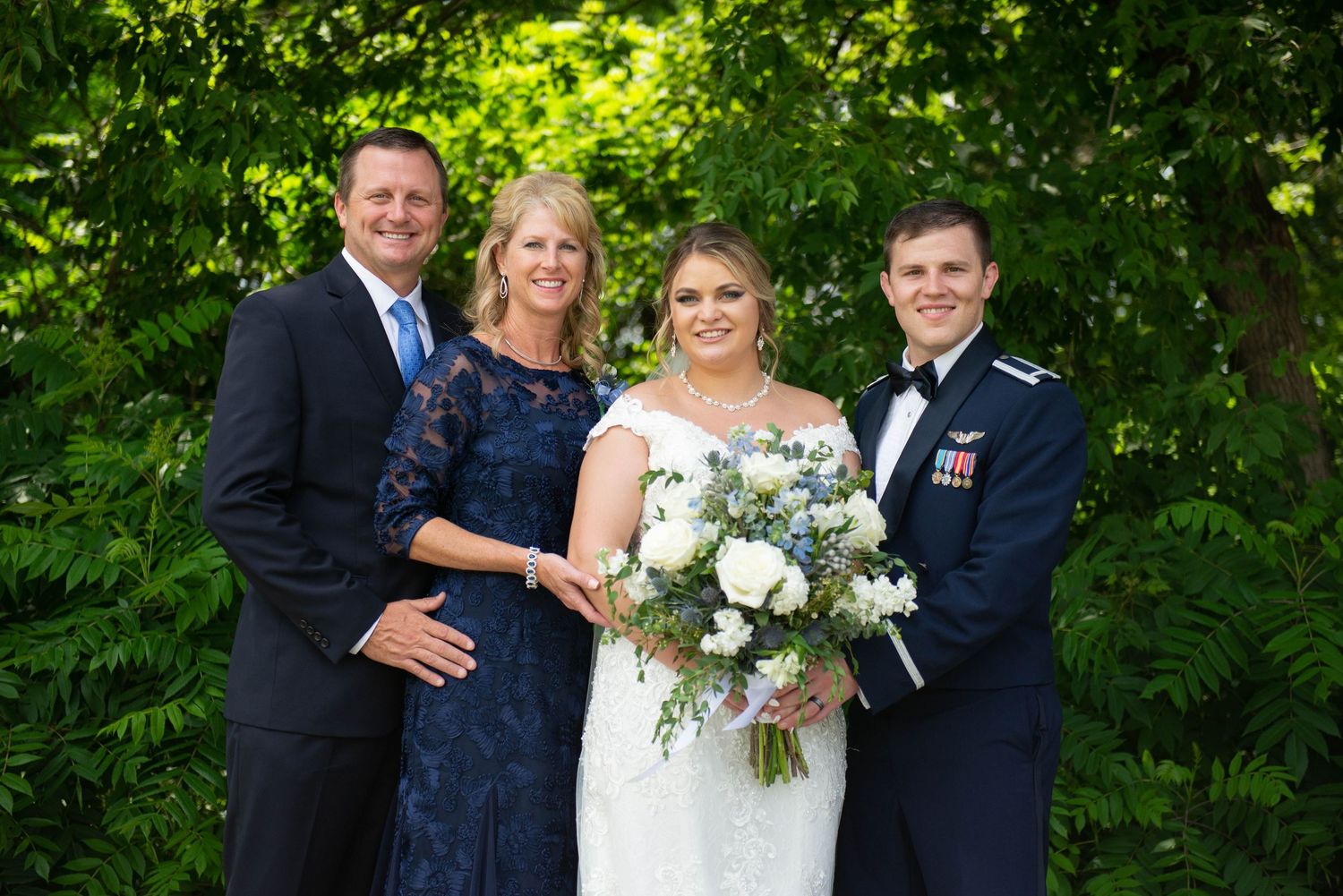 Wedding party posing outdoors against a vibrant green garden backdrop holding white floral bouquets. Zimmerman MN Weddings