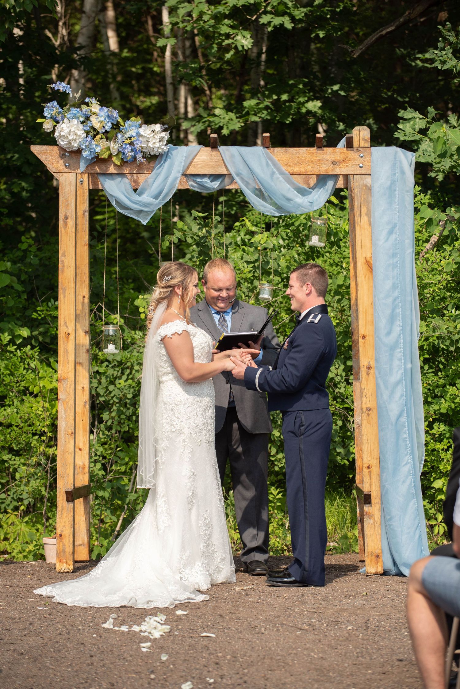An outdoor wedding ceremony takes place under a wooden arch decorated with light blue fabric and floral arrangements. Zimmerman MN Weddings