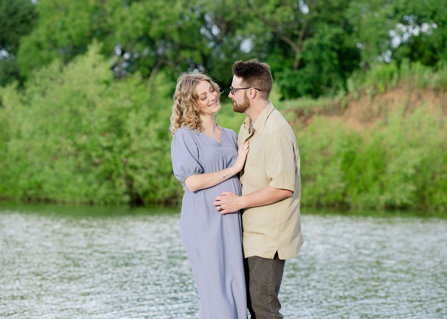 A romantic maternity photoshoot by a lake with a couple embracing in soft light surrounded by lush greenery. Princeton MN Milestone, Materni