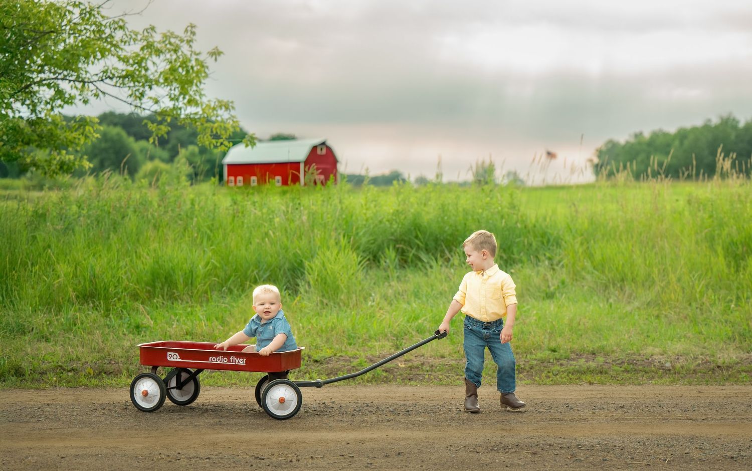 A red barn stands in the background of a green rural field with children playing with a little red wagon.