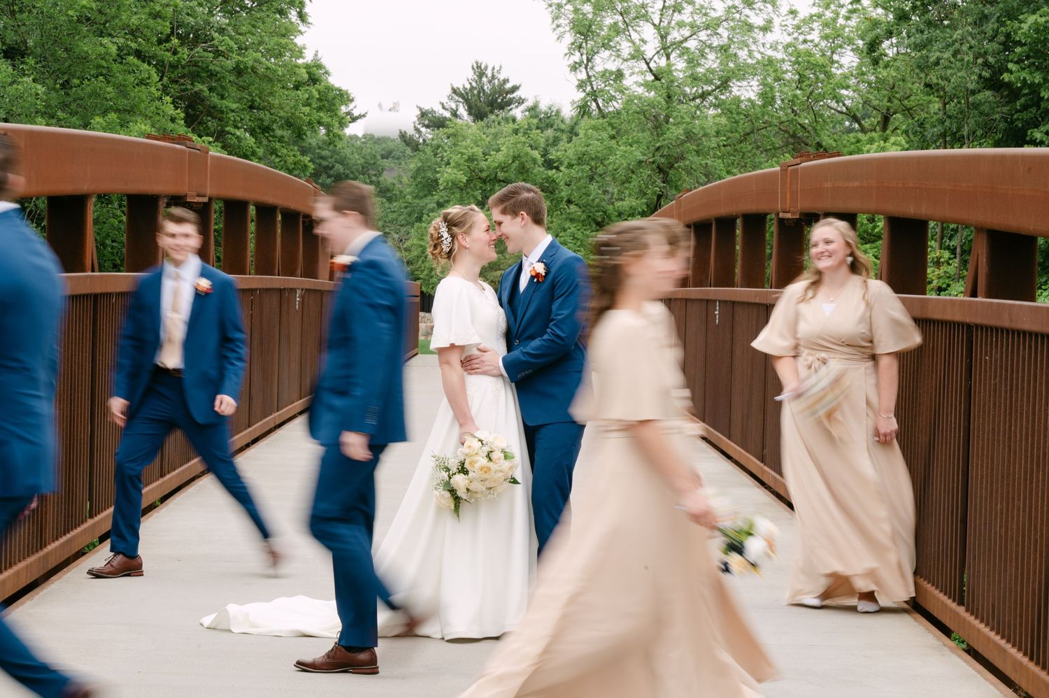 Wedding party poses on a wooden bridge surrounded by lush green trees.