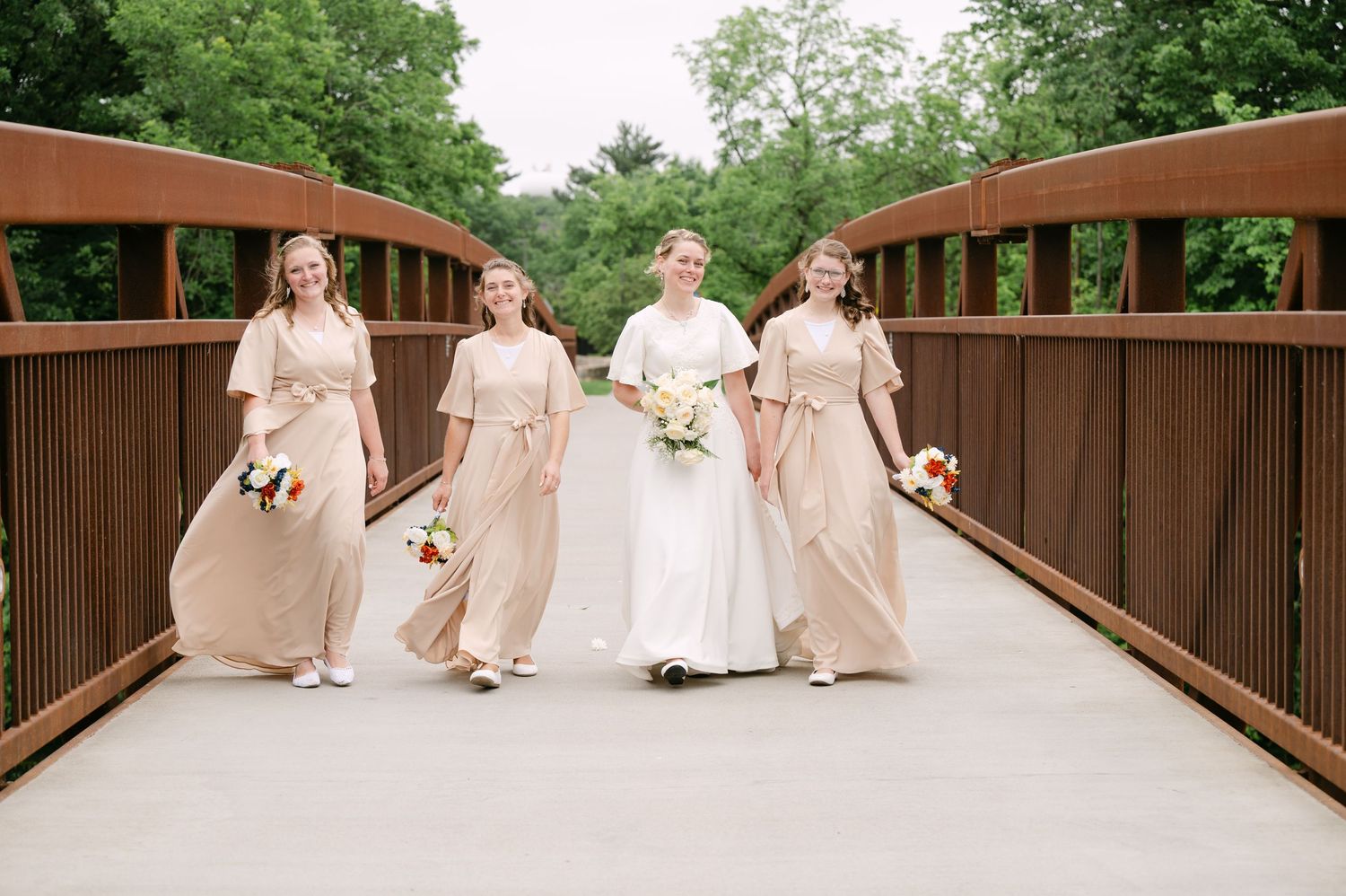 Wedding party in blush pink dresses walks across a wooden bridge during an outdoor ceremony.