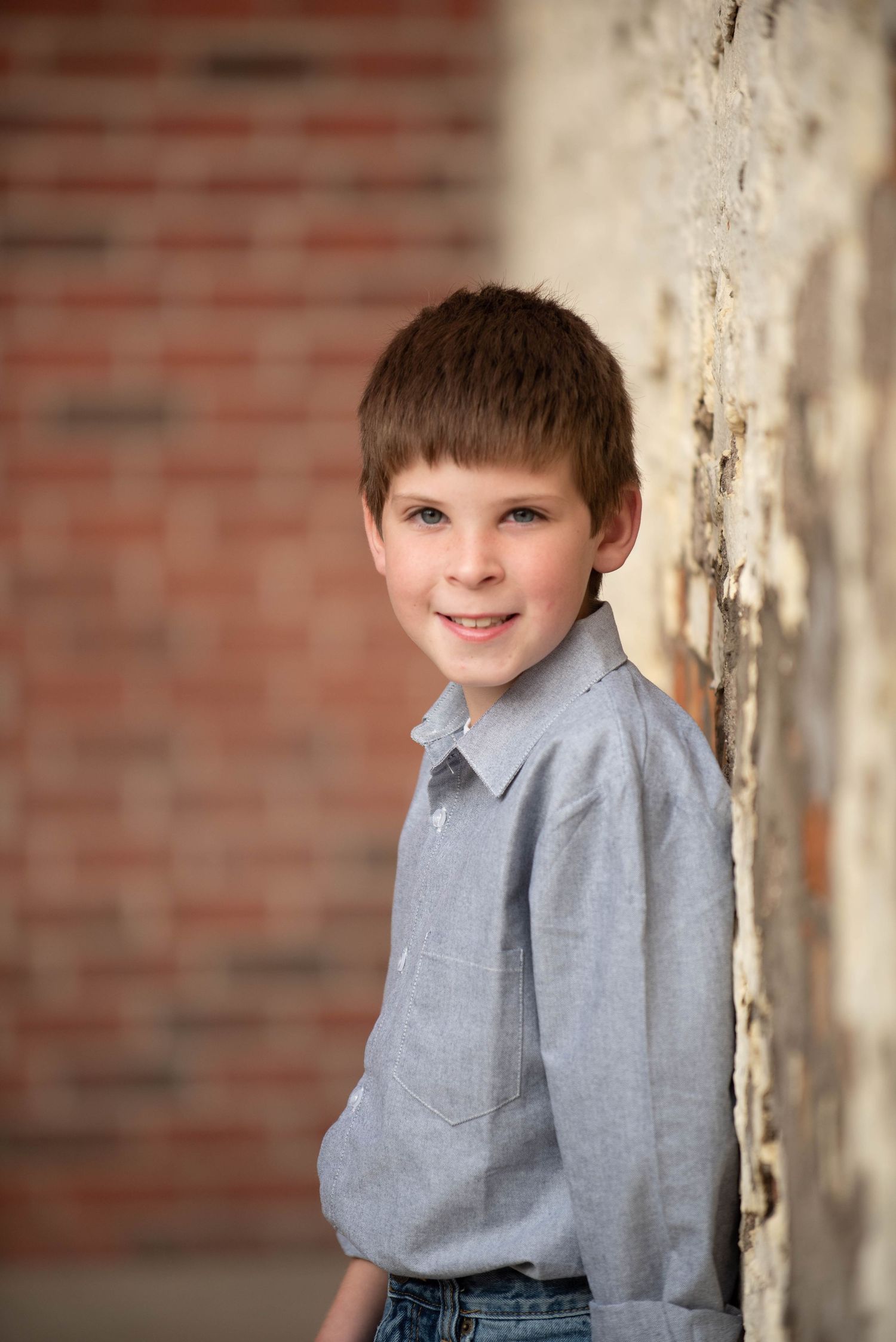 Child wearing a gray button-up shirt poses against a weathered white wall with brick background.