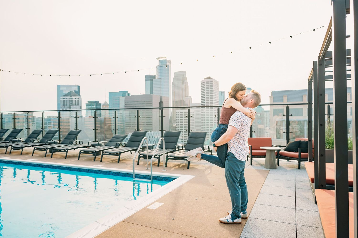 A rooftop pool deck with lounge chairs and string lights overlooking a city skyline.