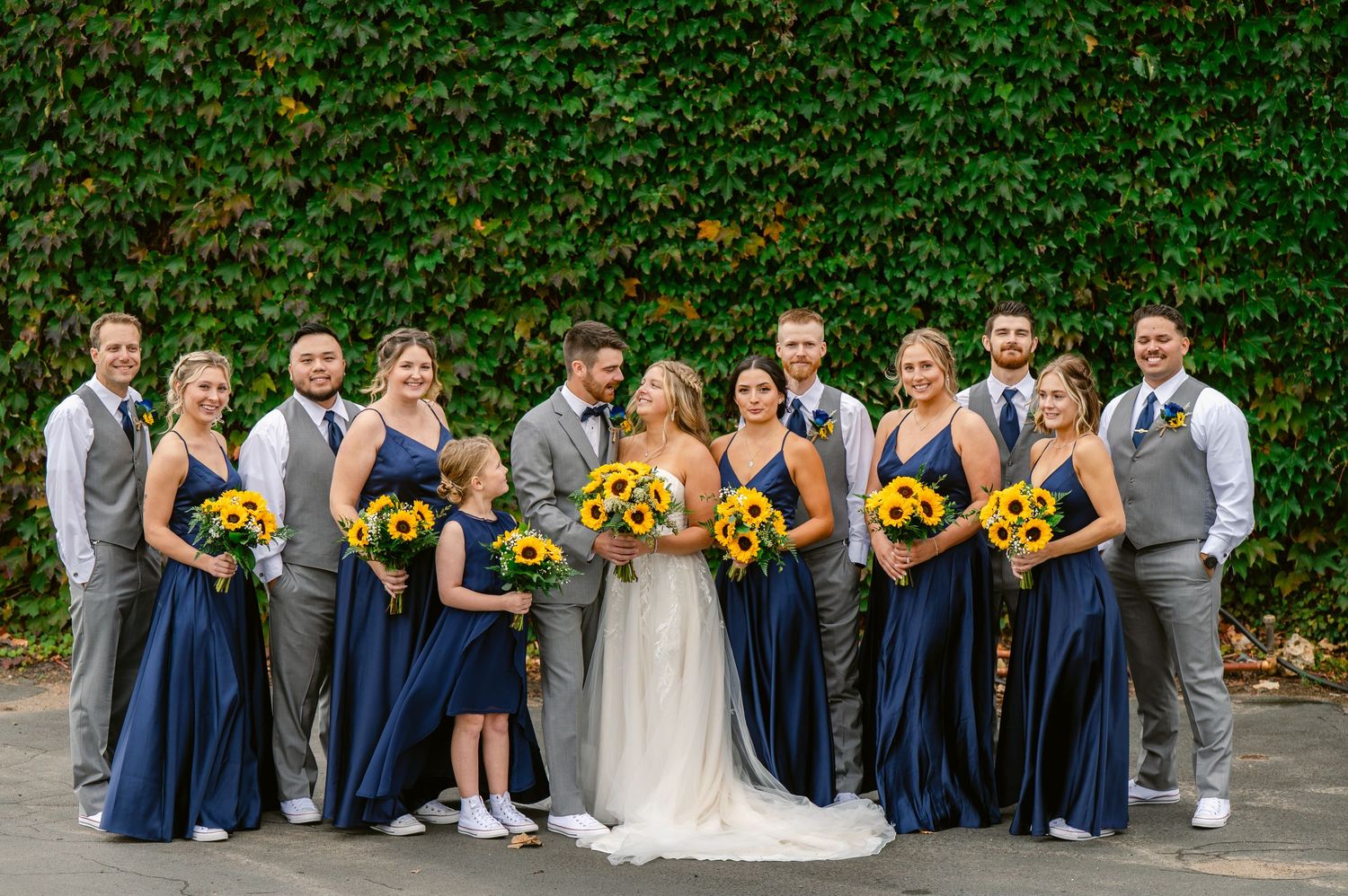 Central MN Wedding Photos | Wedding party in navy blue and gray posing with sunflower bouquets by ivy wall.