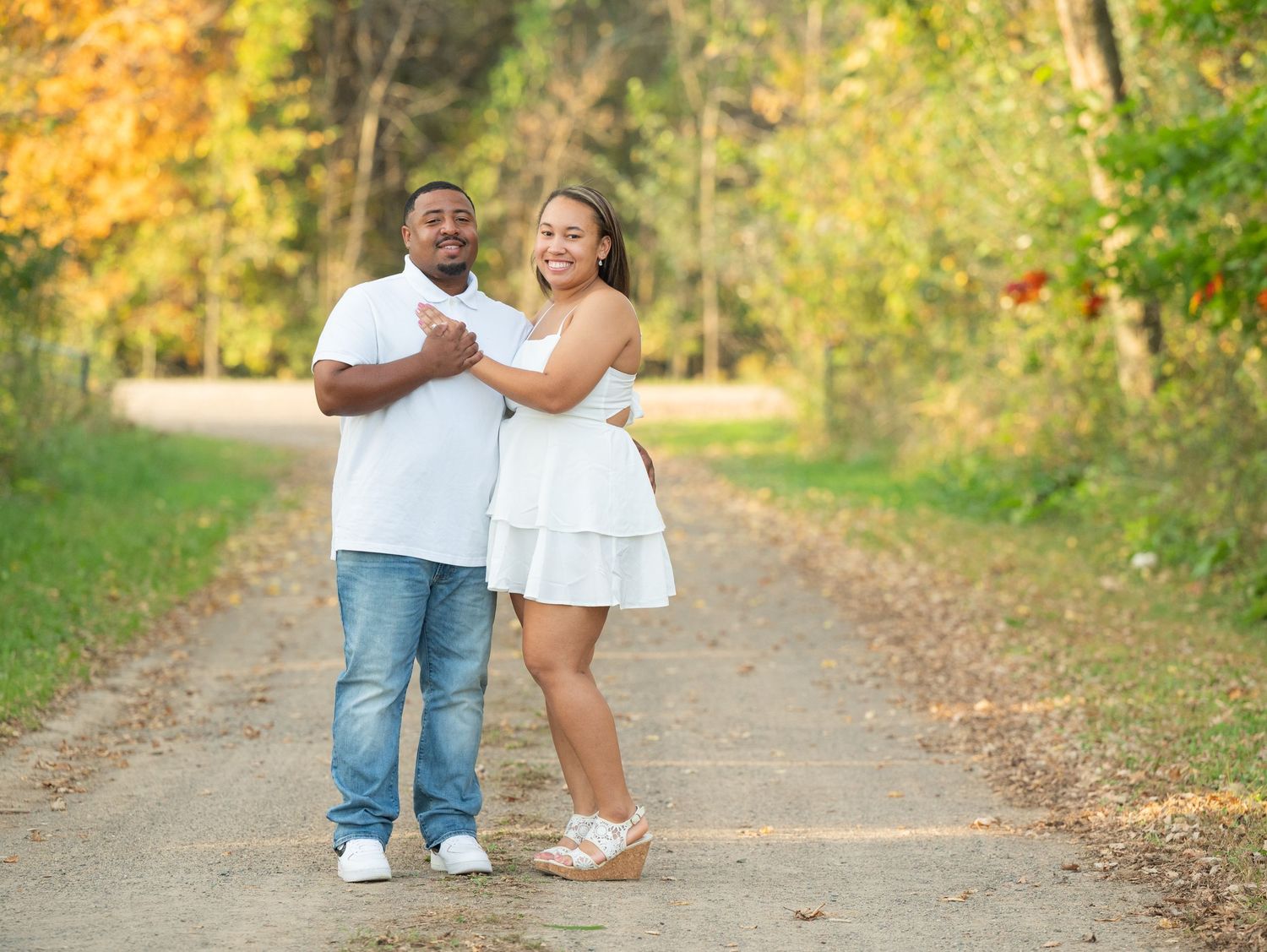 Central Minnesota Best Engagement Photographer - A couple in white outfits poses happily on a tree-lined path surrounded by autumn foliage.