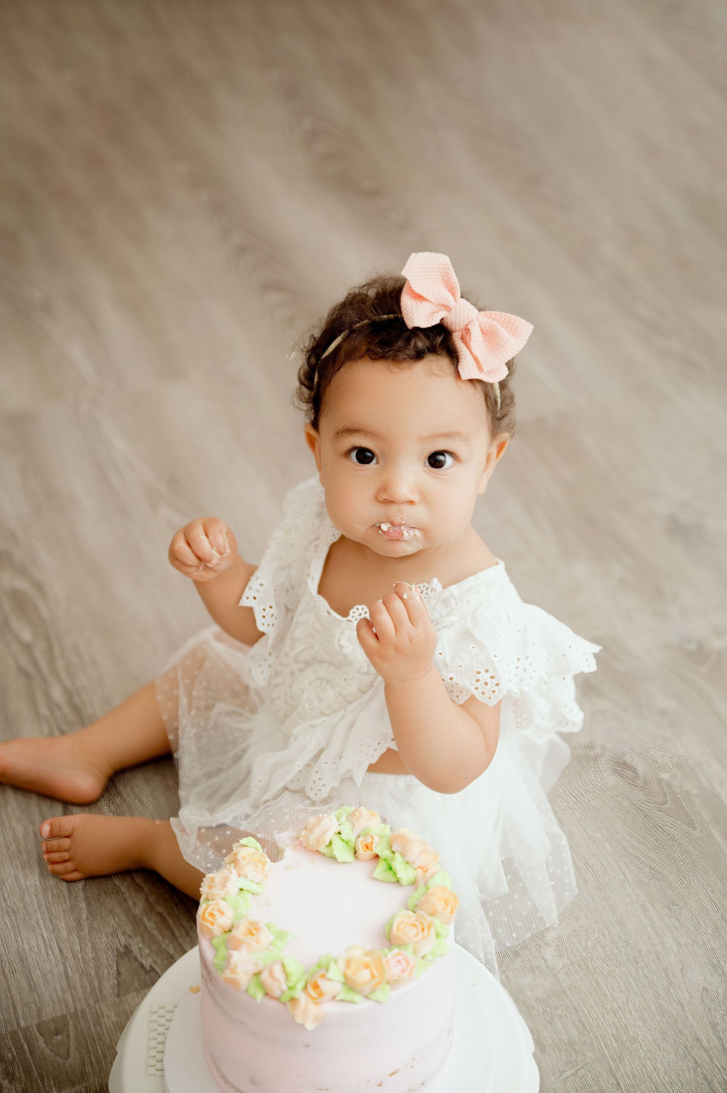 Central MN Milestone Photos - Baby girl in white lace dress with pink bow smashes first birthday cake on studio floor.