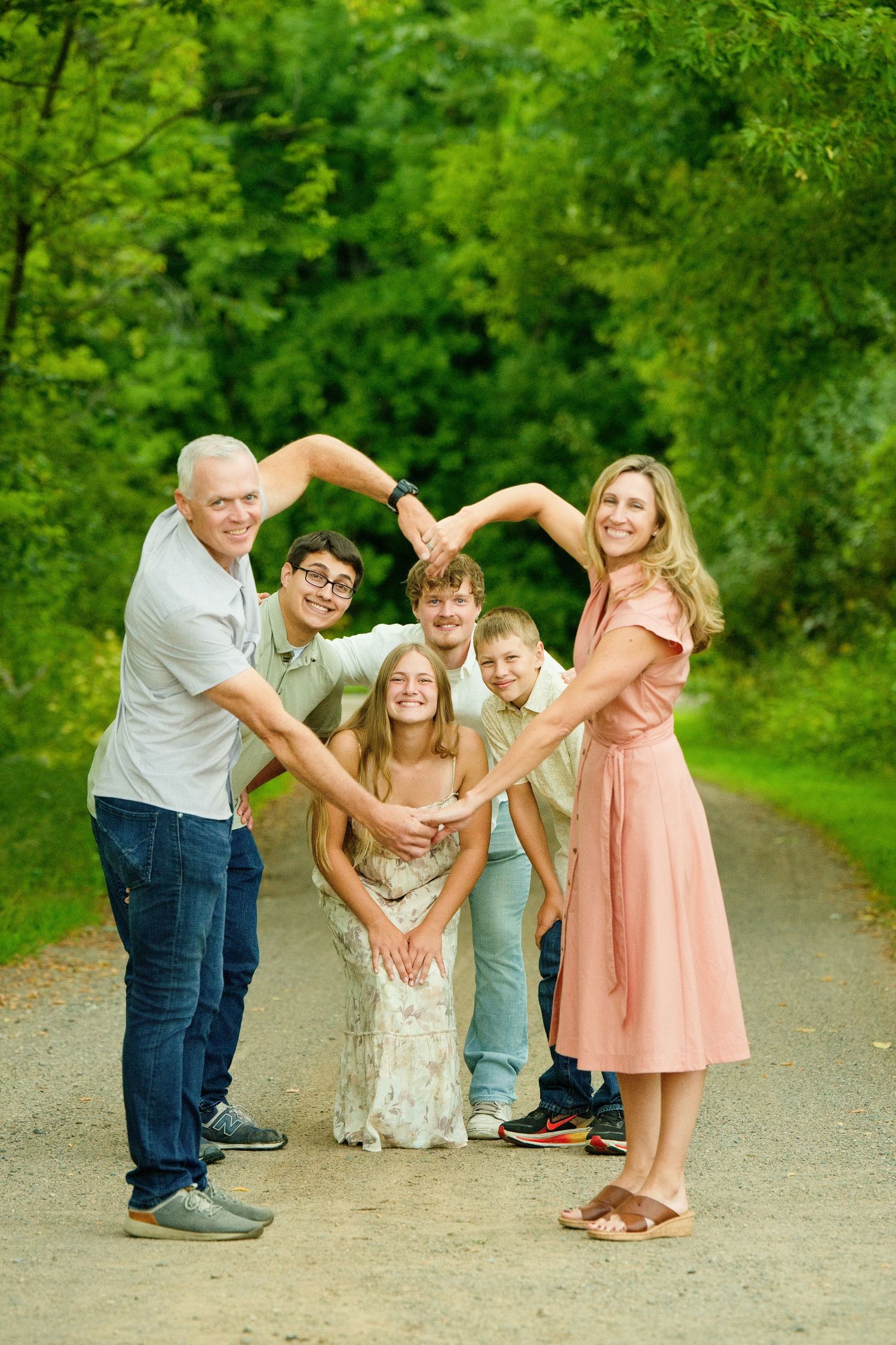 A happy family of five forms a heart shape with their arms on a tree-lined path outdoors.