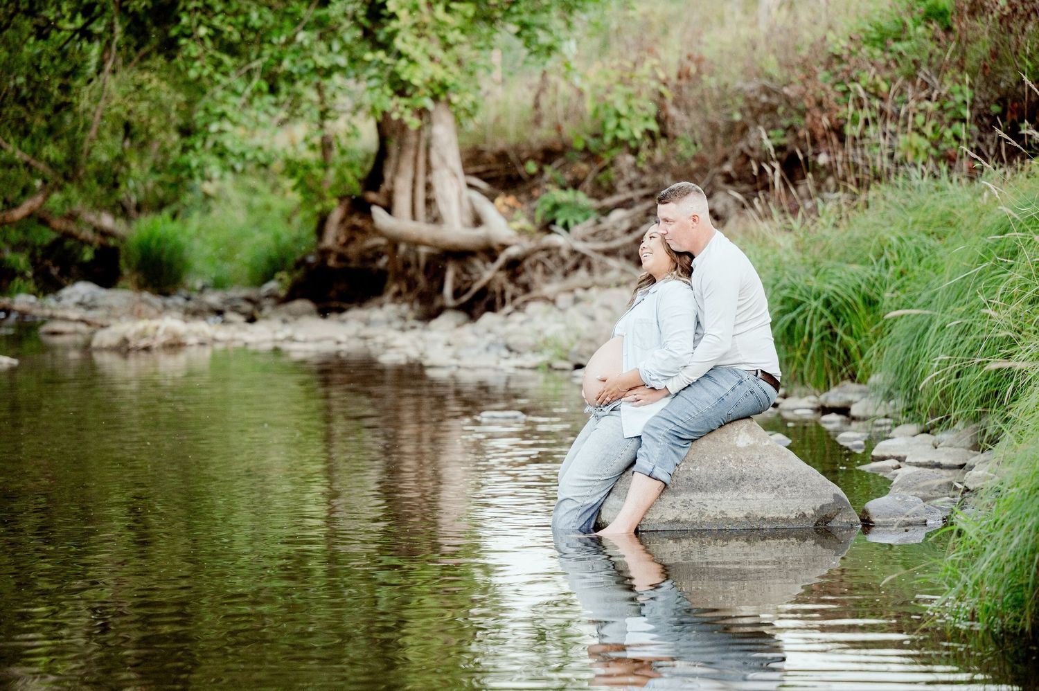 A relaxed person in light summer attire sits at the edge of a tranquil pond surrounded by lush greenery and plants.