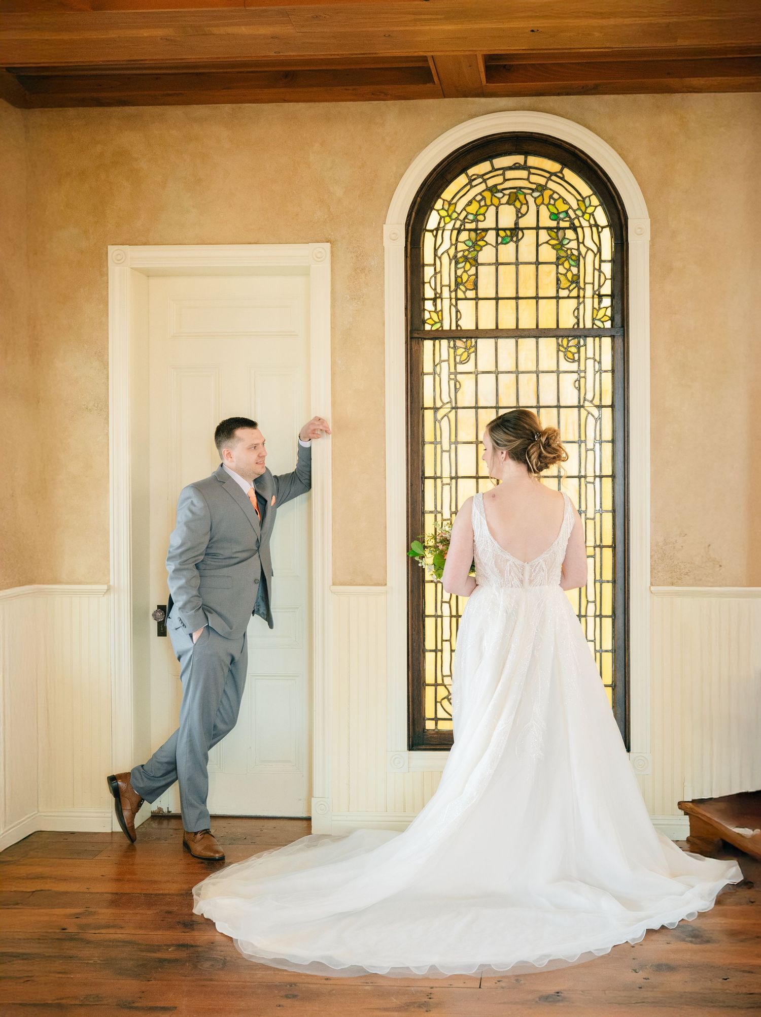 Bride in flowing white gown and groom in gray suit share first look near stained glass window.