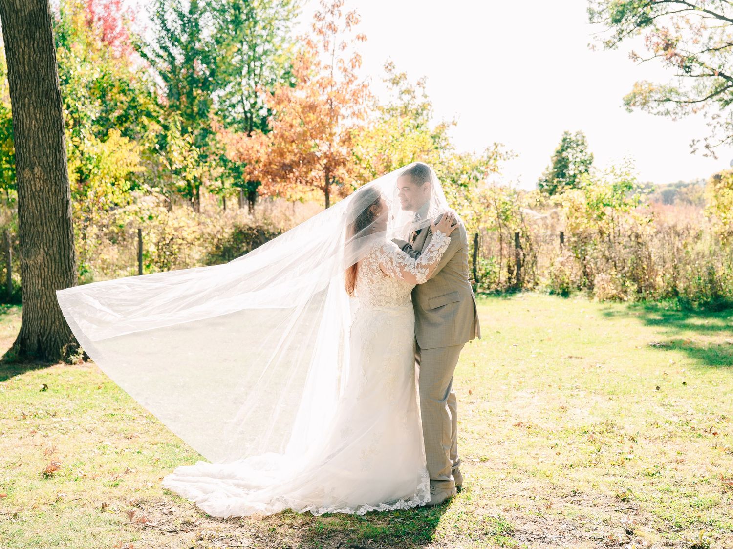 A bride and groom share a kiss outdoors in autumn, her white veil flowing in the breeze amid colorful fall foliage.