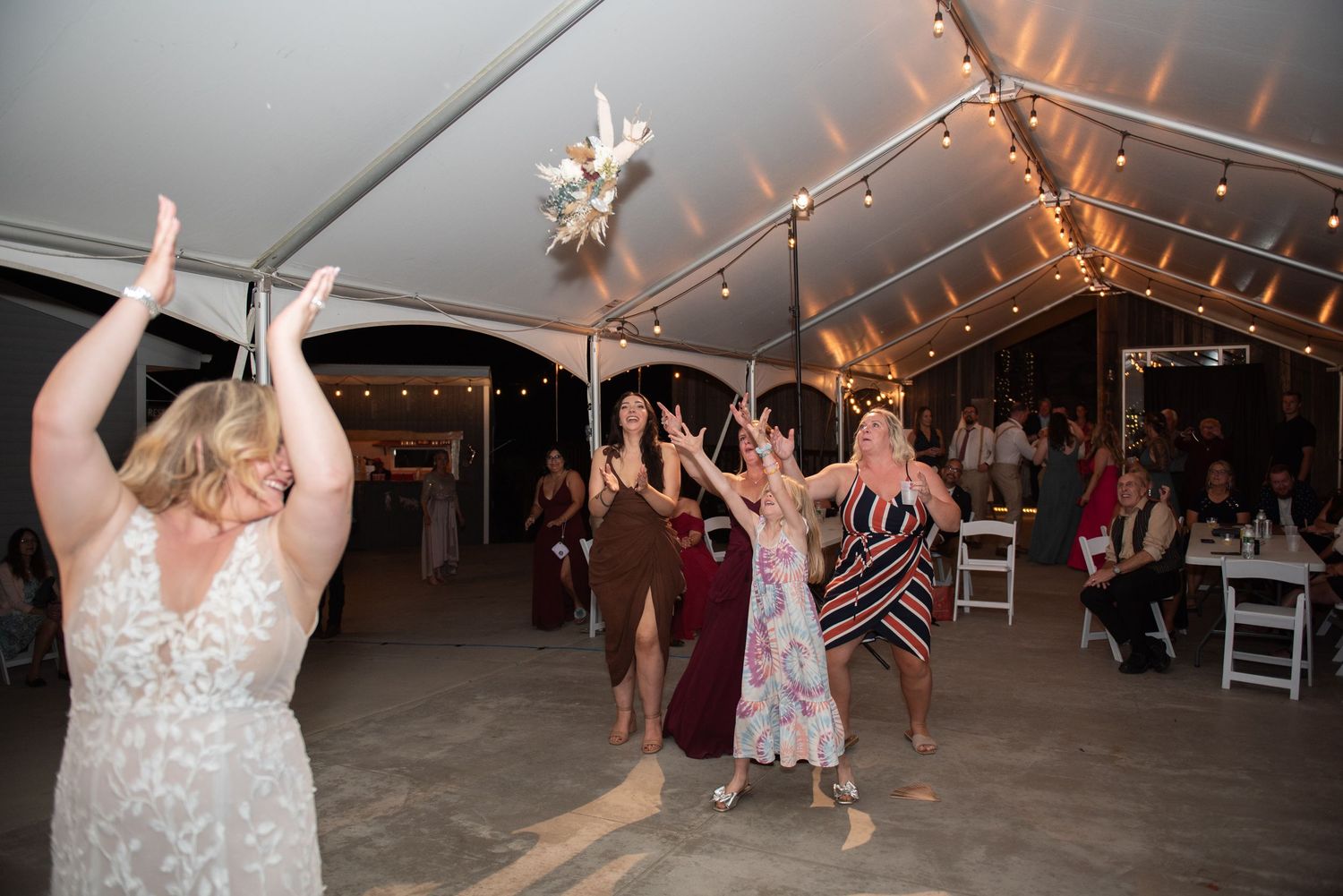 Wedding guests dance and celebrate under a tent with twinkling lights at a reception.