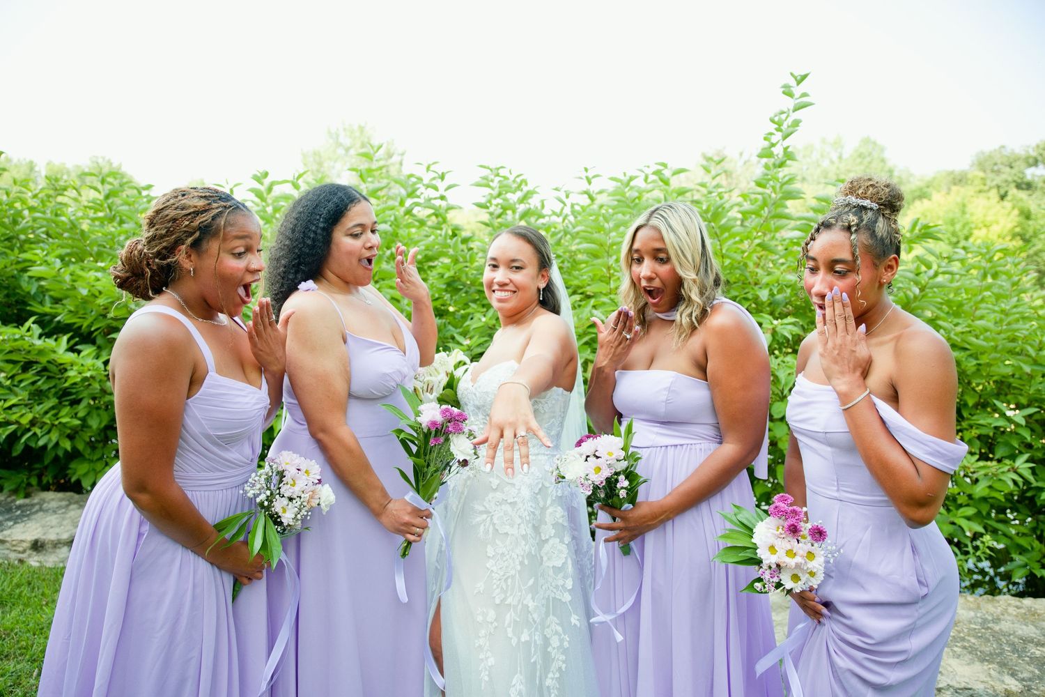 Bride in floral gown shows ring to four bridesmaids in lavender dresses holding bouquets outdoors.