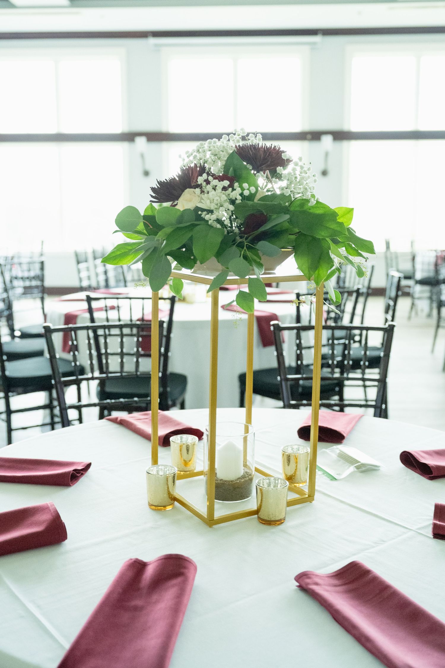 Elegant wedding reception table with gold geometric centerpiece, burgundy napkins, white tablecloth, and floral arrangement.
