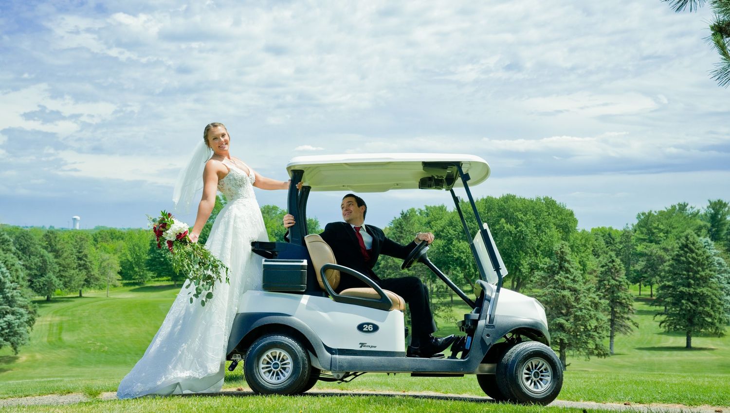 Bride in white gown holding bouquet poses beside golf cart on lush green course.