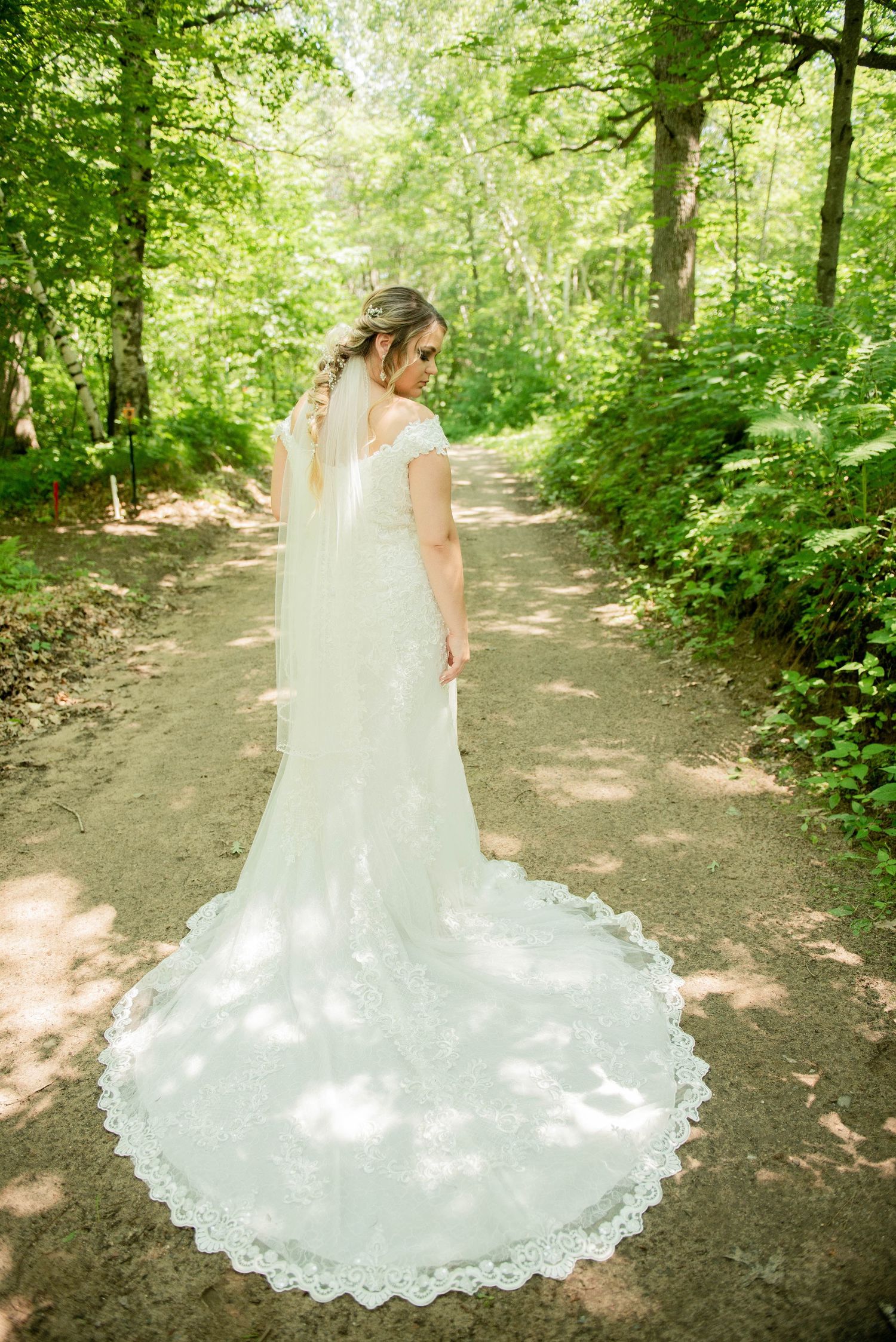 A lace wedding dress with long train photographed from behind on a sunlit forest path. Zimmerman MN Weddings