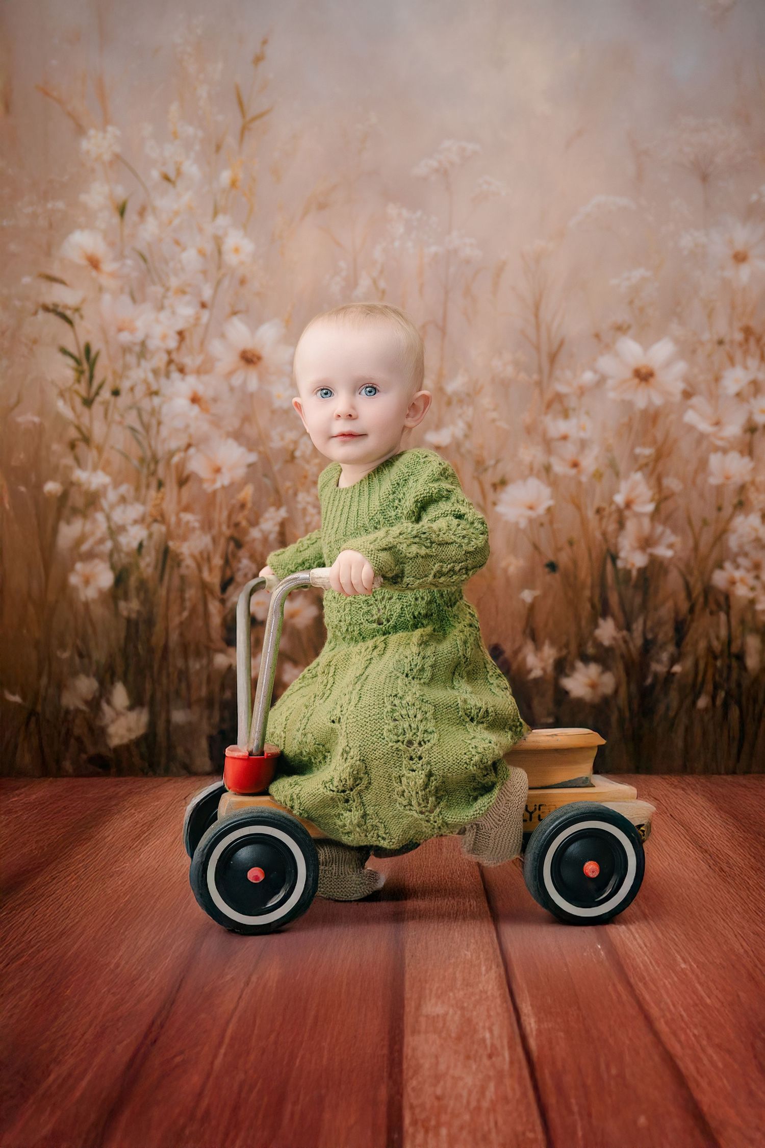 Baby girl in green knit dress riding a wooden toy tricycle on red wood floor with floral backdrop. mother day photoshoot