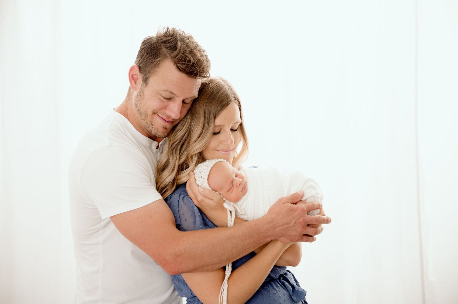 A couple in white shirts tenderly holds their newborn baby against a soft white background.
