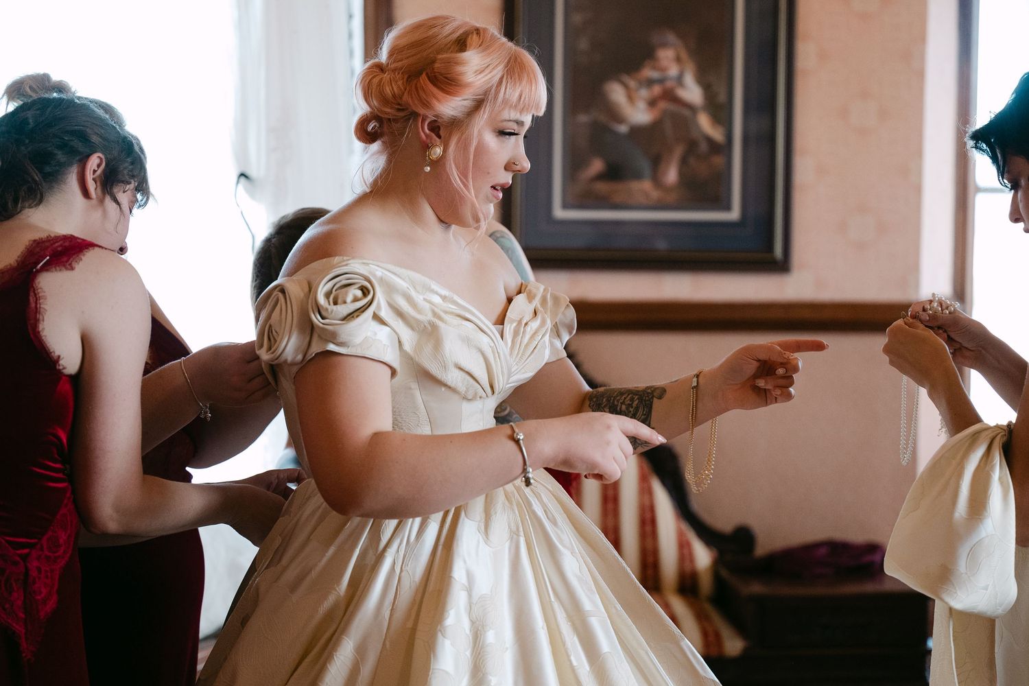 Central MN Wedding | Bride in ivory off-shoulder gown being assisted by bridesmaids in a vintage-styled room during wedding preparations.