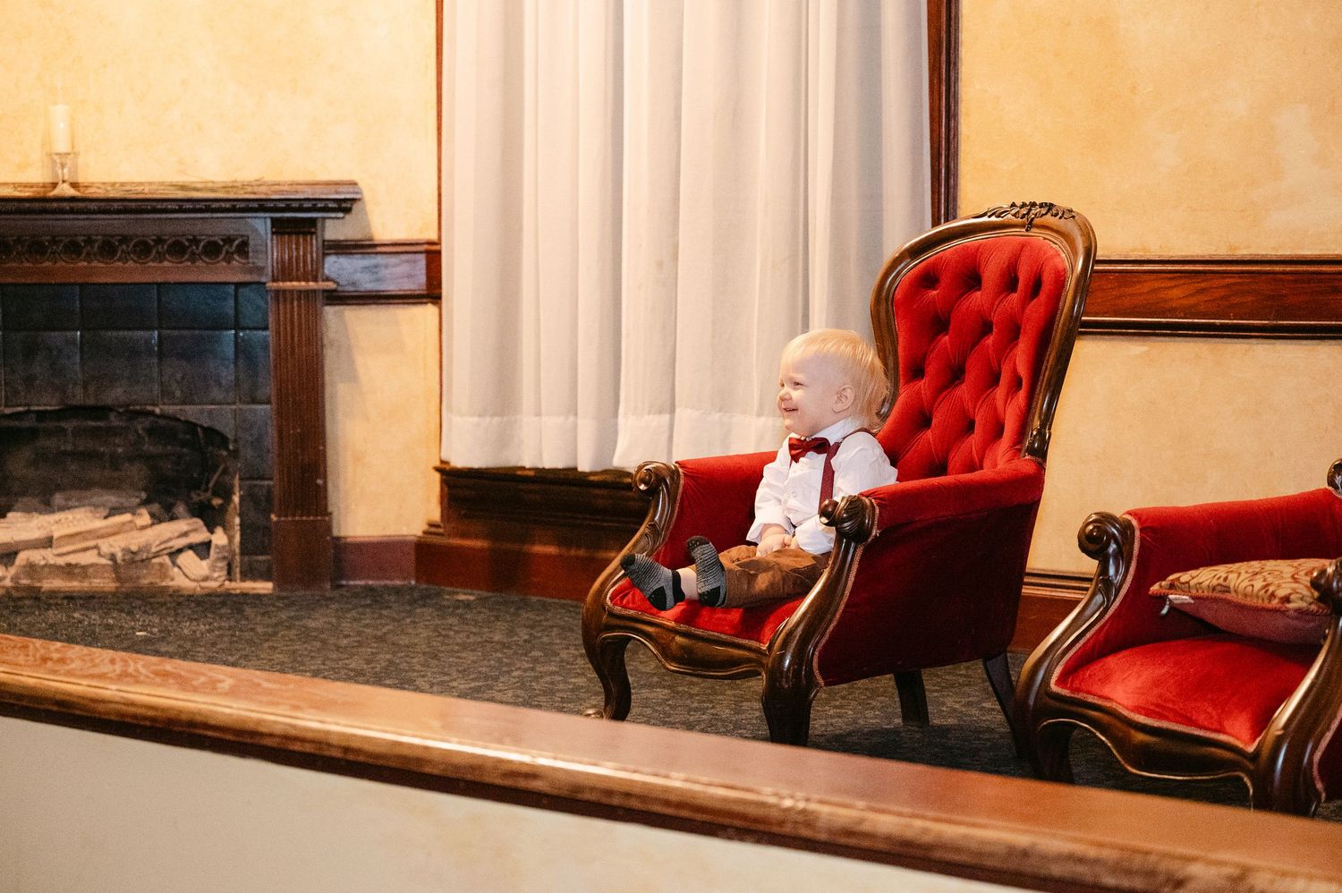 Central MN Wedding Photos | Adorable baby in white outfit sitting in ornate red velvet chair near fireplace.