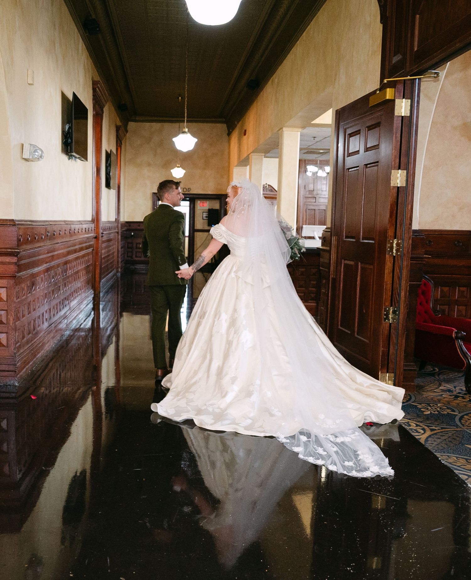 Central MN Wedding Photos | Bride in voluminous white gown and veil walks with groom through ornate historic venue hallway.