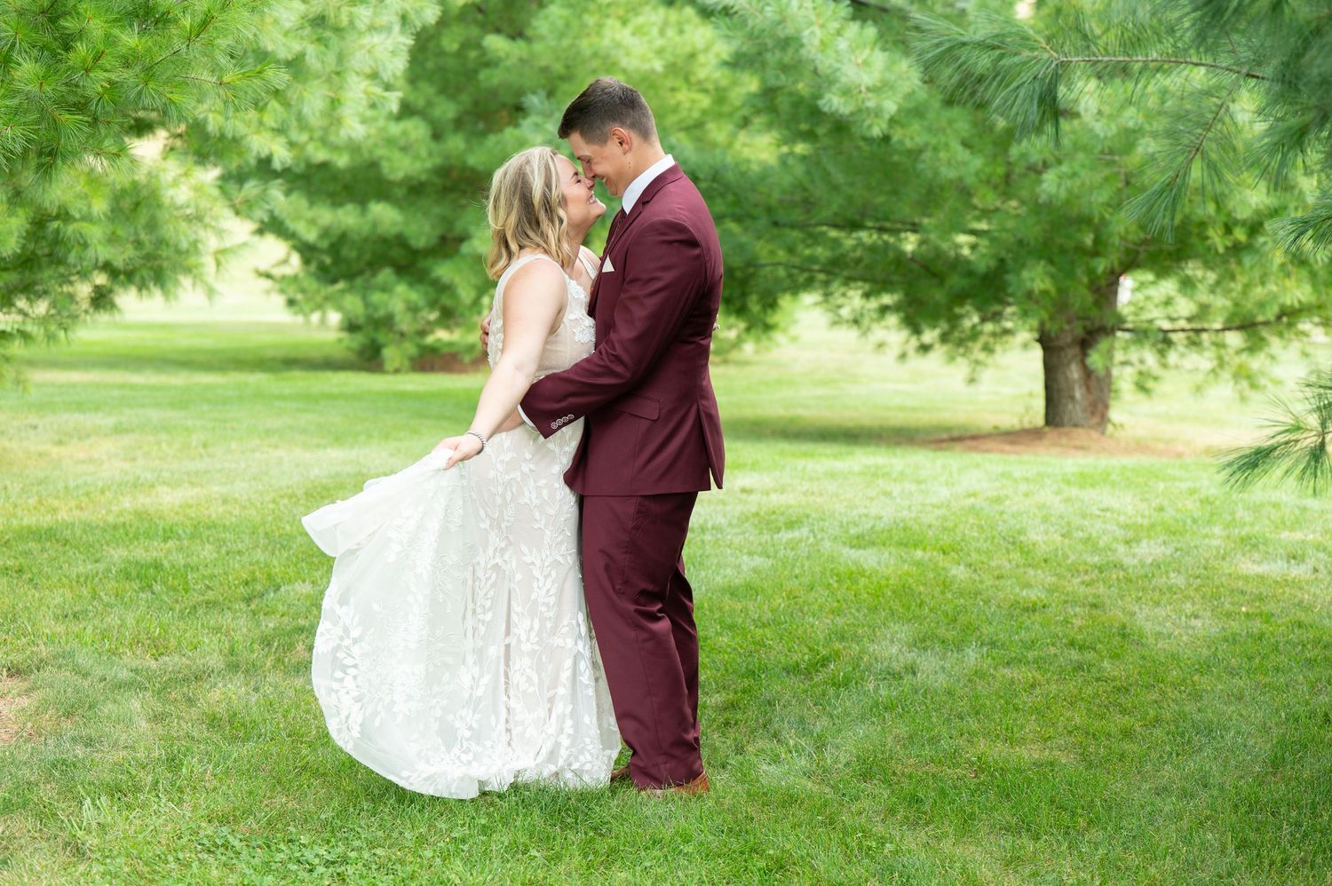 Central MN Wedding Photography - Bride and groom share a romantic moment in lush green park, she in lace gown, he in burgundy suit.