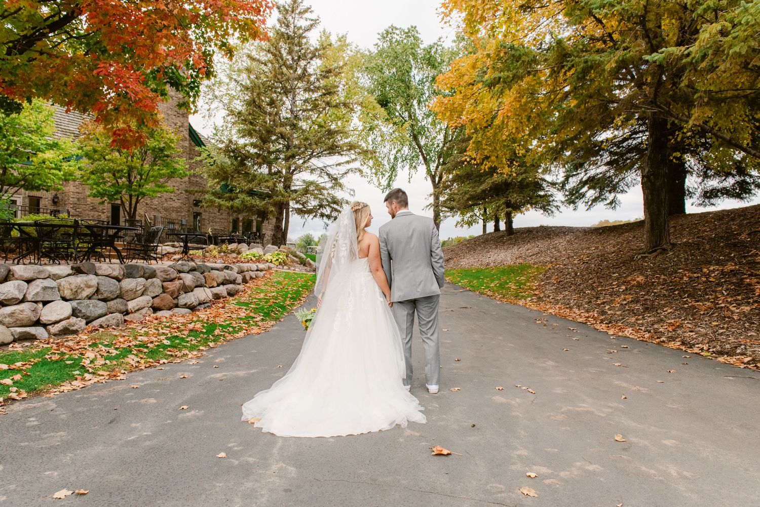 Central MN Wedding Photos | Bride and groom walk hand-in-hand down a tree-lined path surrounded by colorful fall foliage.