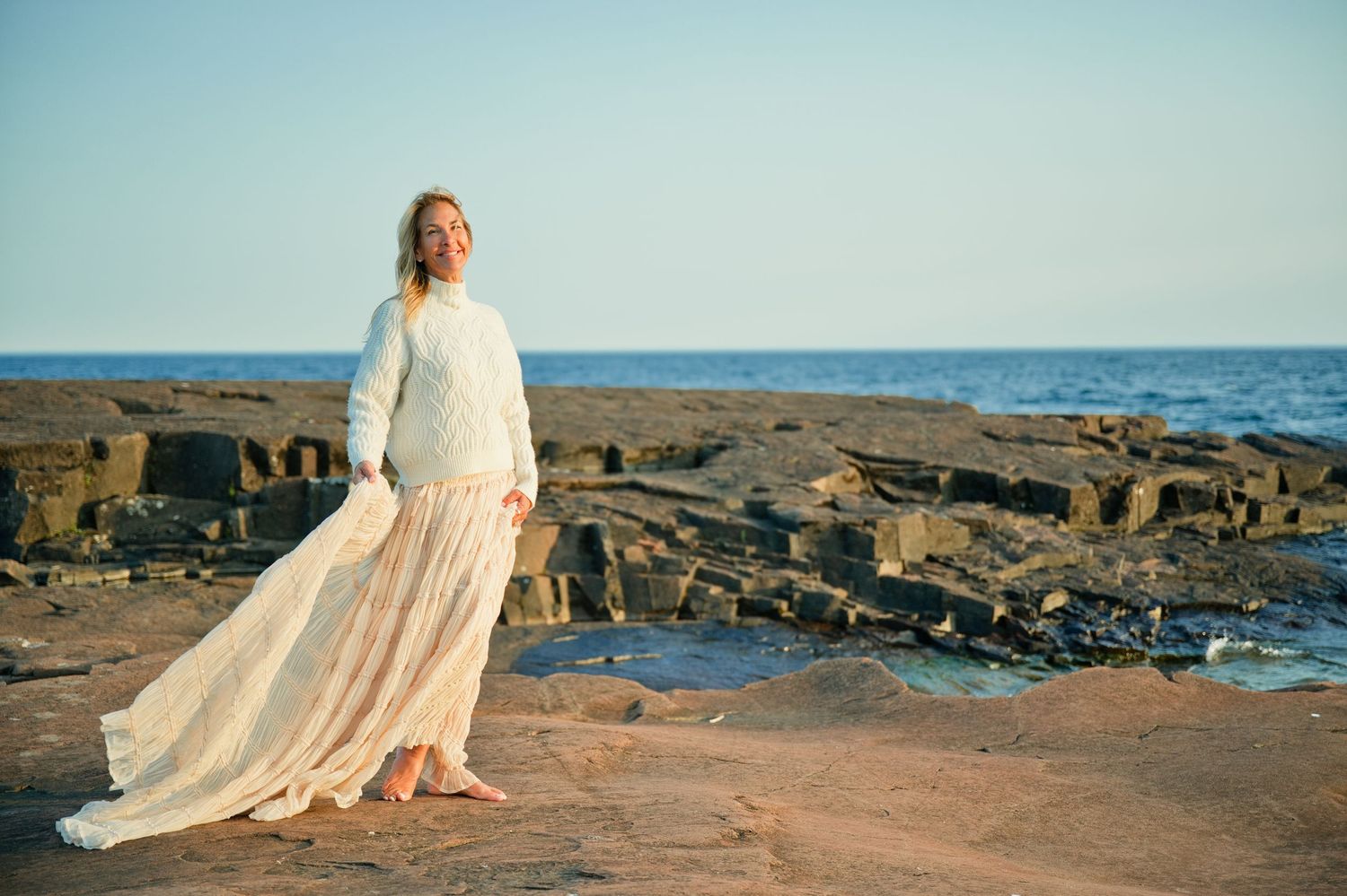 Woman in white sweater and flowing skirt stands on rocky coastal shore with ocean waves in golden sunlight.