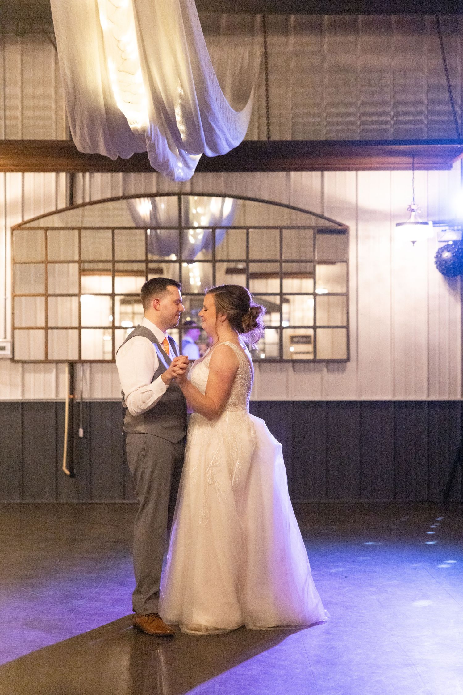 Bride and groom share their first dance under soft purple lighting in a rustic barn venue.