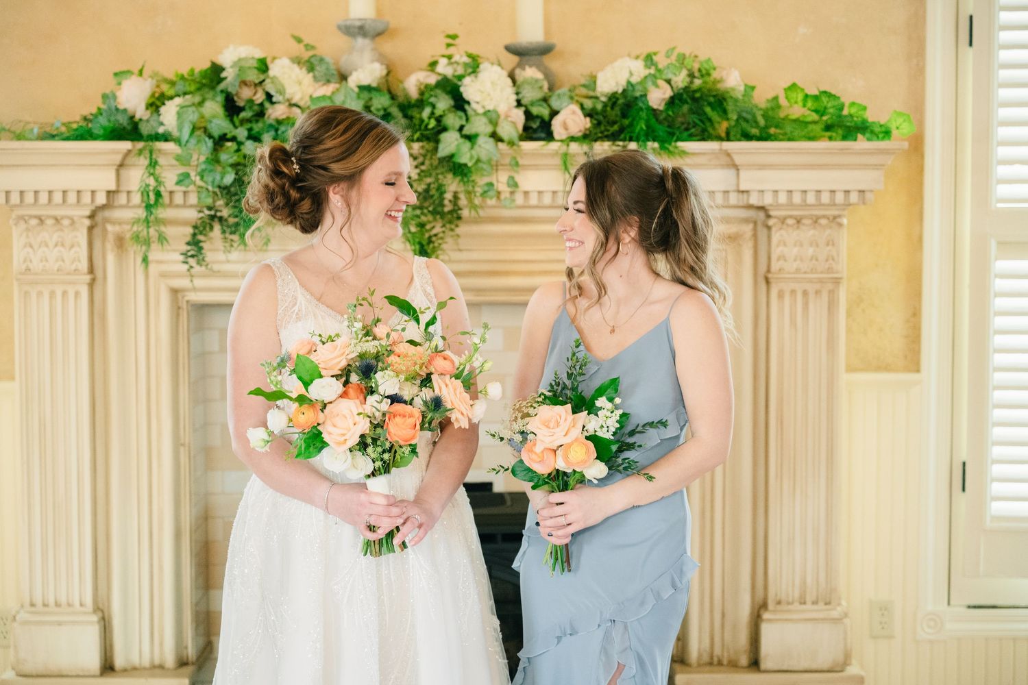 Bride and bridesmaid in blue dress share a smile holding peach floral bouquets.