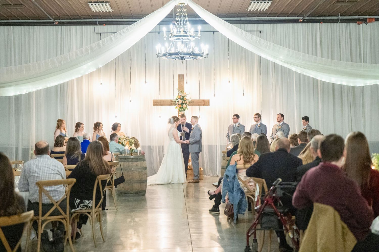 Wedding ceremony with guests seated as couple stands at altar beneath chandelier and white draped ceiling.