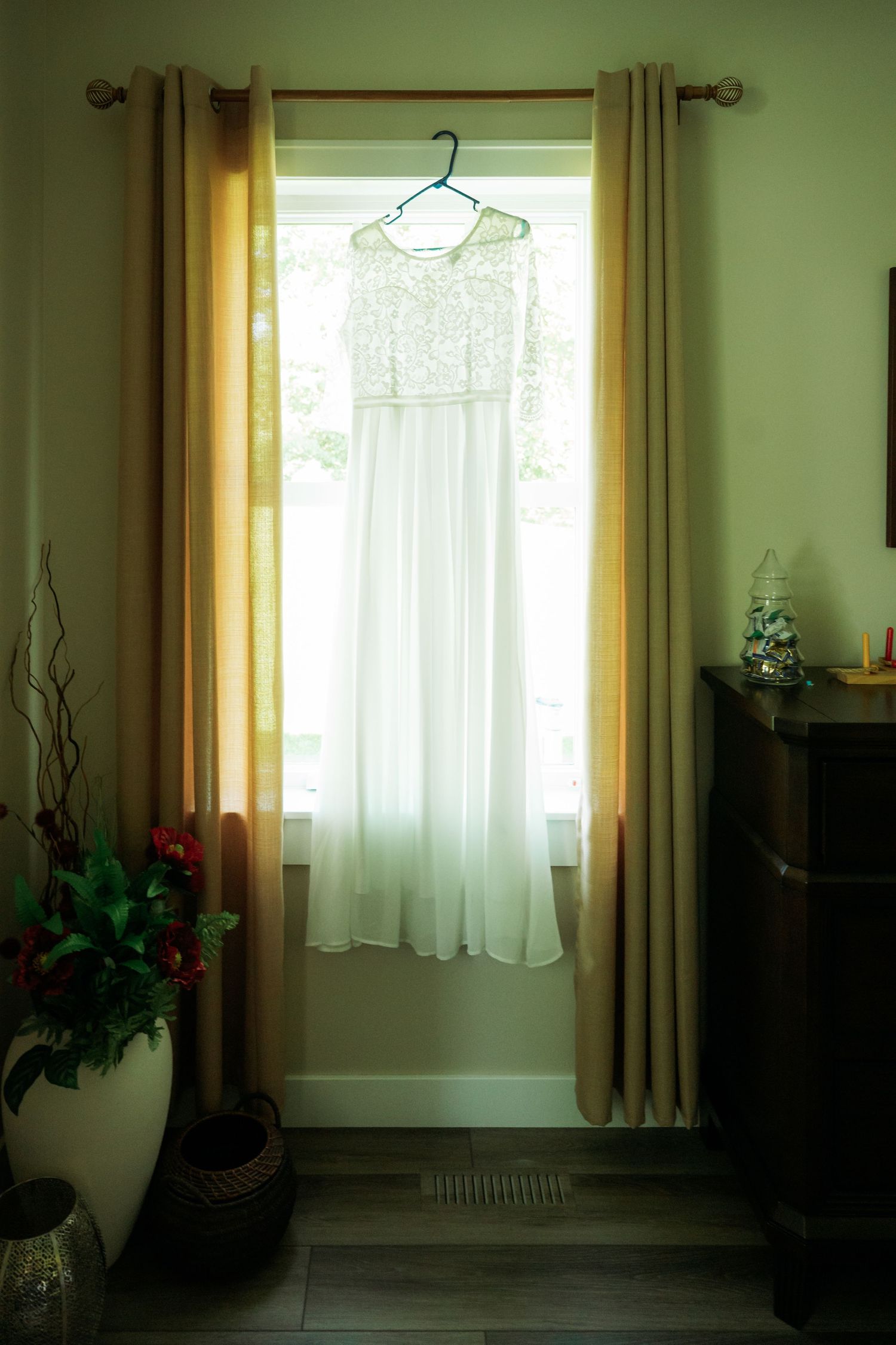 White lace wedding dress hanging on a hanger in a sunlit window with golden curtains and hardwood floors.