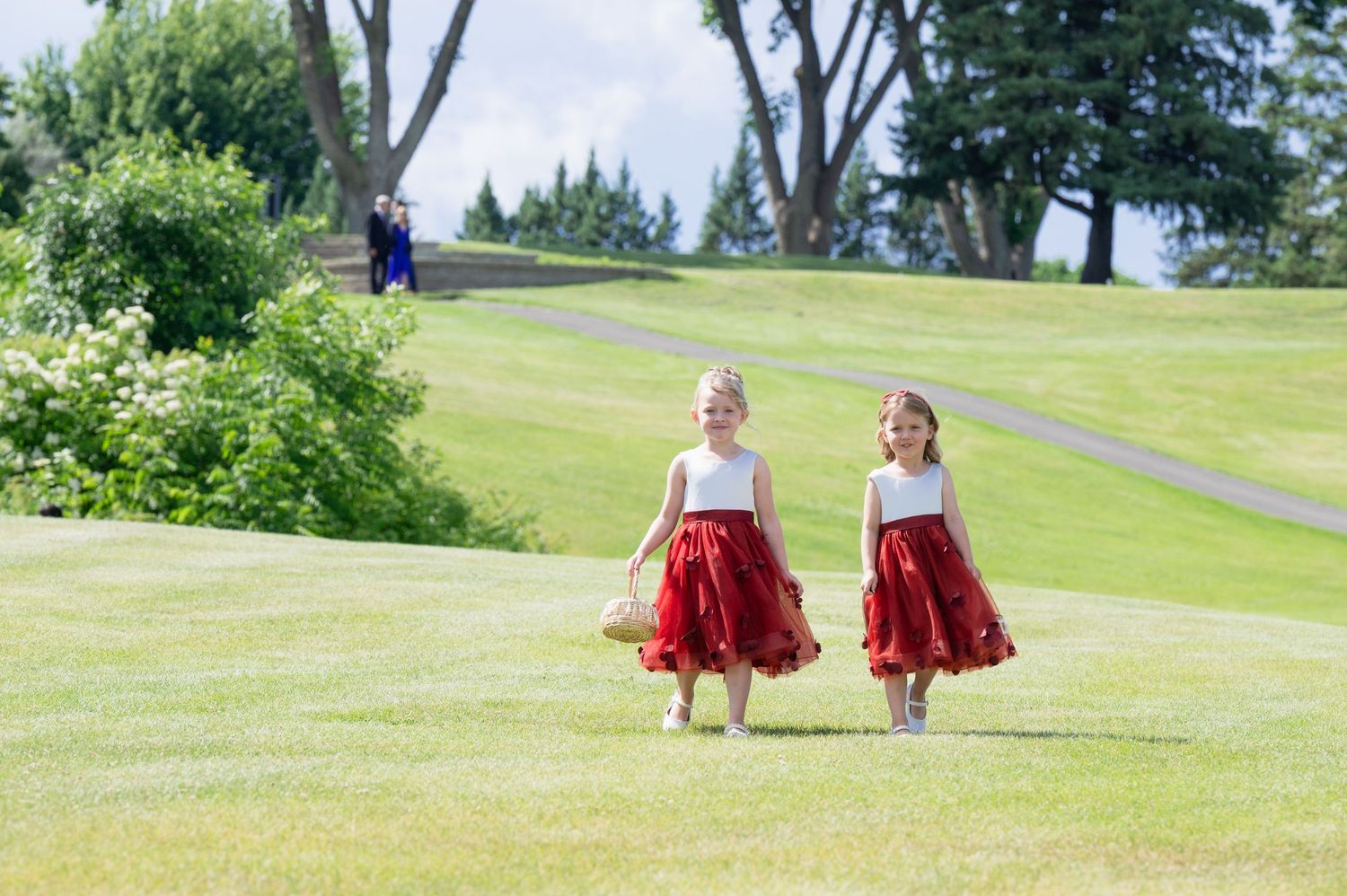 Two young flower girls in white and red dresses walk across a lush green golf course during a wedding.