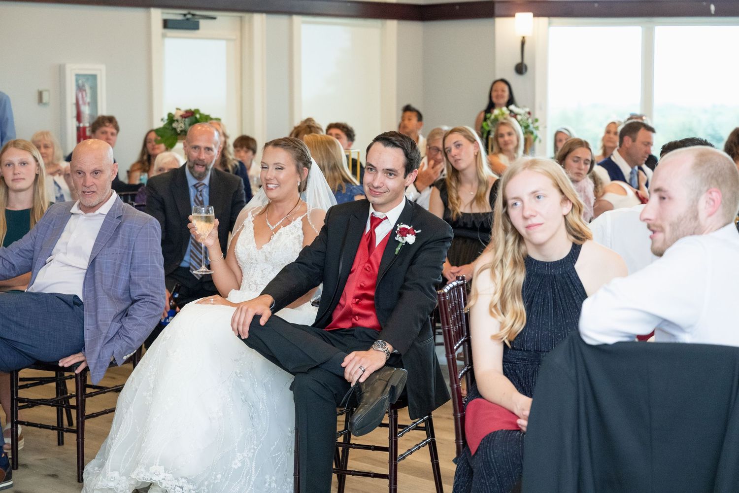 Bride and groom sit with guests during an indoor wedding ceremony, groom wearing a red tie.