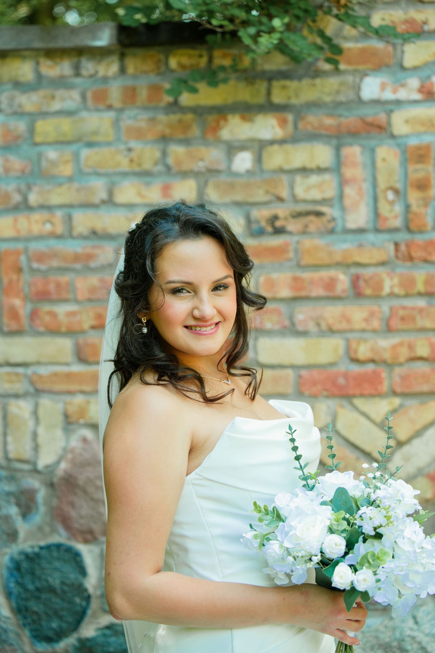 Smiling bride in strapless white gown holding bouquet of white flowers against rustic brick wall backdrop.