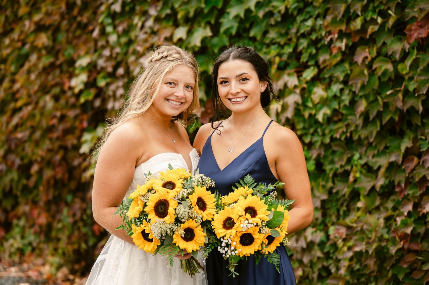 Central MN Wedding Photos | Bride and bridesmaid smile together holding vibrant sunflower bouquets in front of an autumn ivy wall.