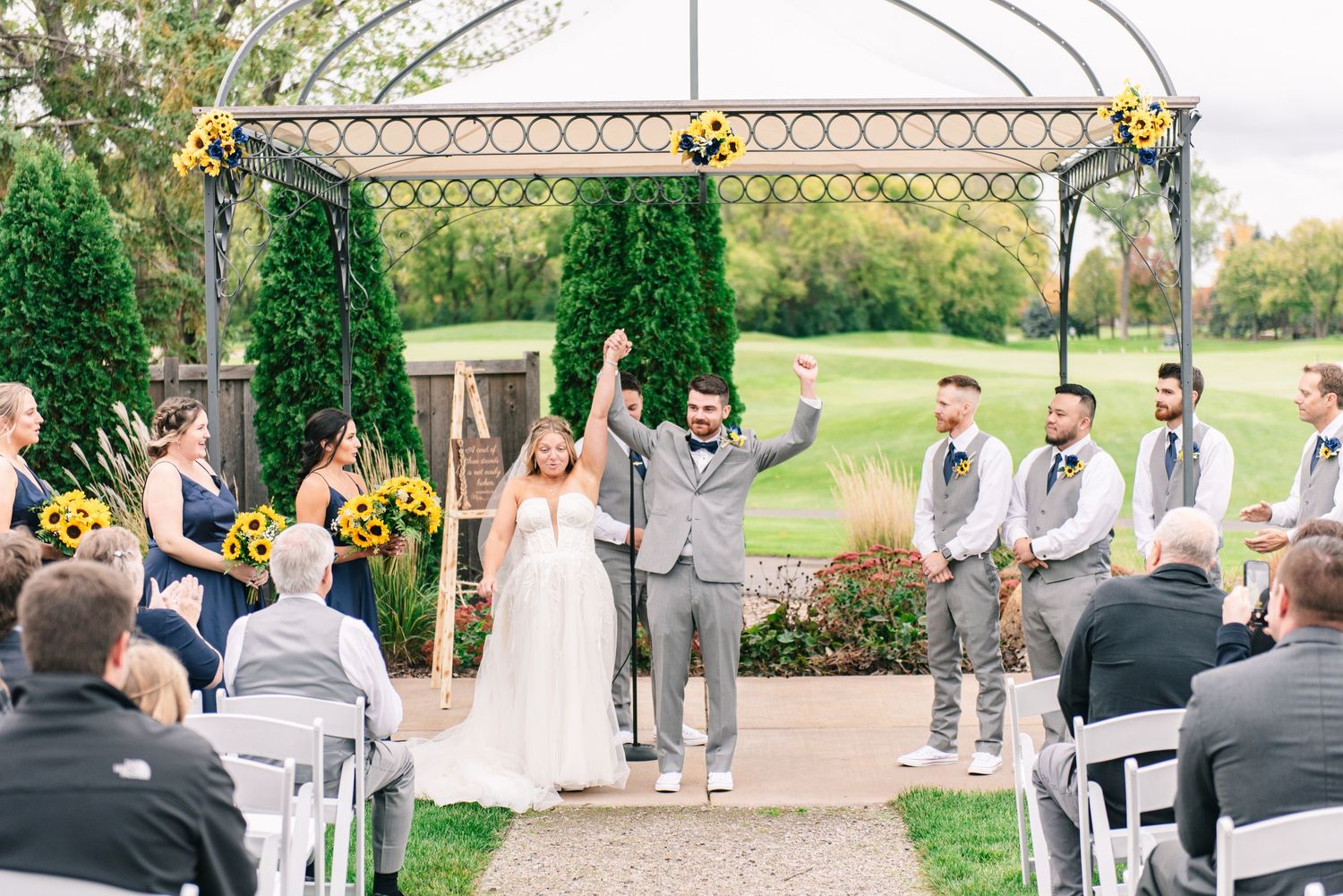 Central MN Wedding Photos - Bride and groom celebrate with raised hands at outdoor wedding ceremony beneath arbor on golf course.