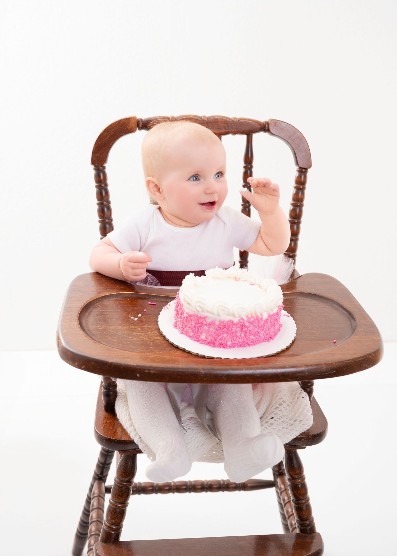 Central MN cake smash Photography - A happy baby in a wooden high chair celebrates their first birthday with a pink and white frosted cake.