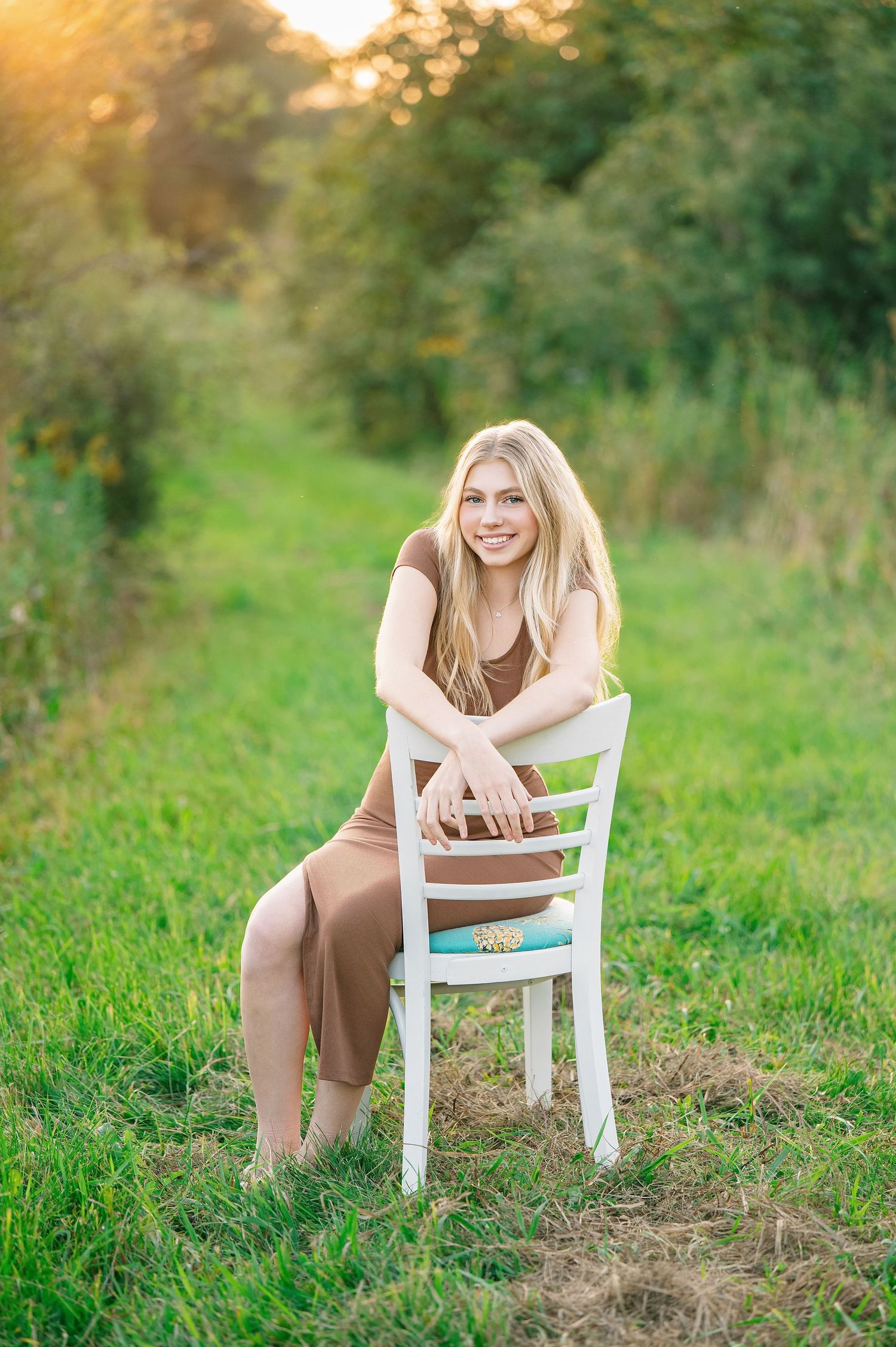 Central MN Senior Photography - Young blonde woman in brown dress sitting backwards on white chair in sunny green meadow, smiling at camera.