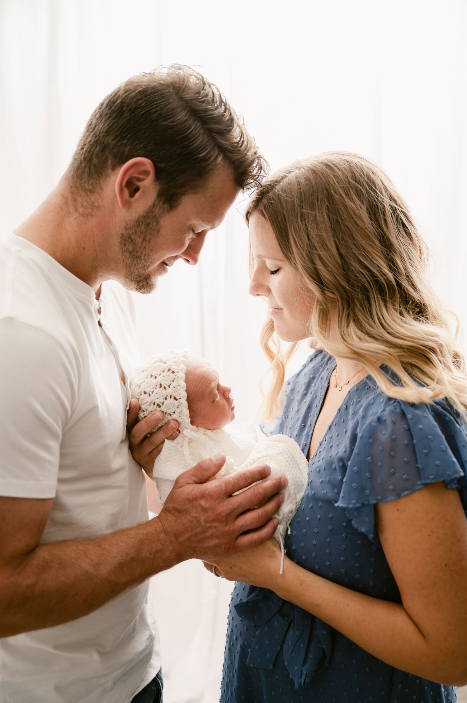 New parents tenderly gaze at their newborn baby wrapped in white lace, sharing a precious family moment.