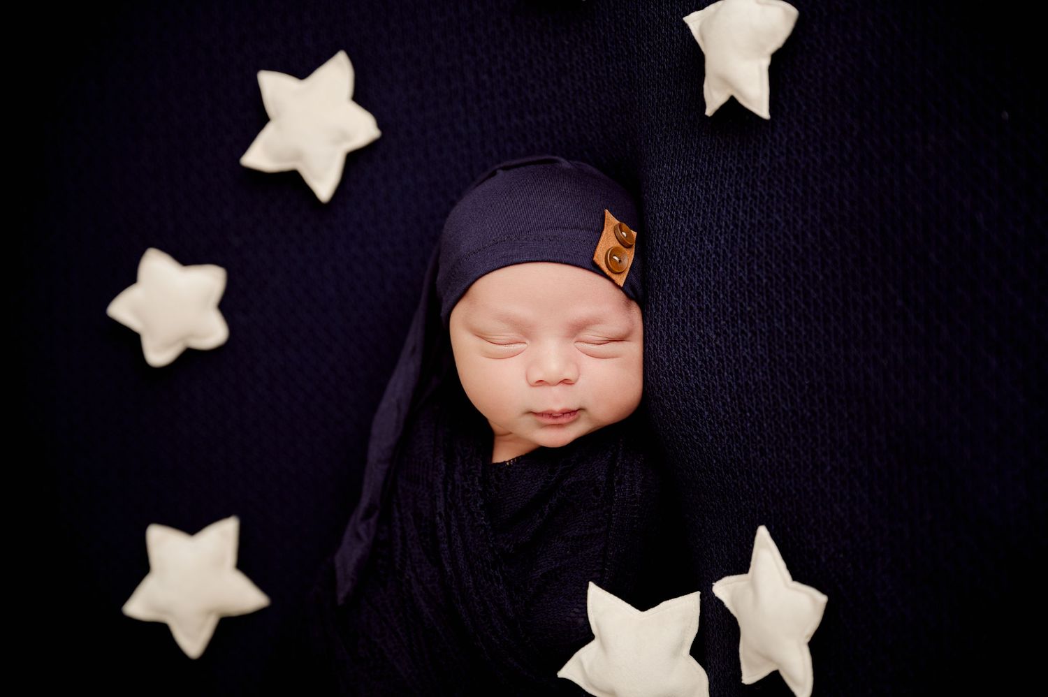 Newborn baby swaddled in navy blue wrap and hat, sleeping peacefully surrounded by white star props on dark background.