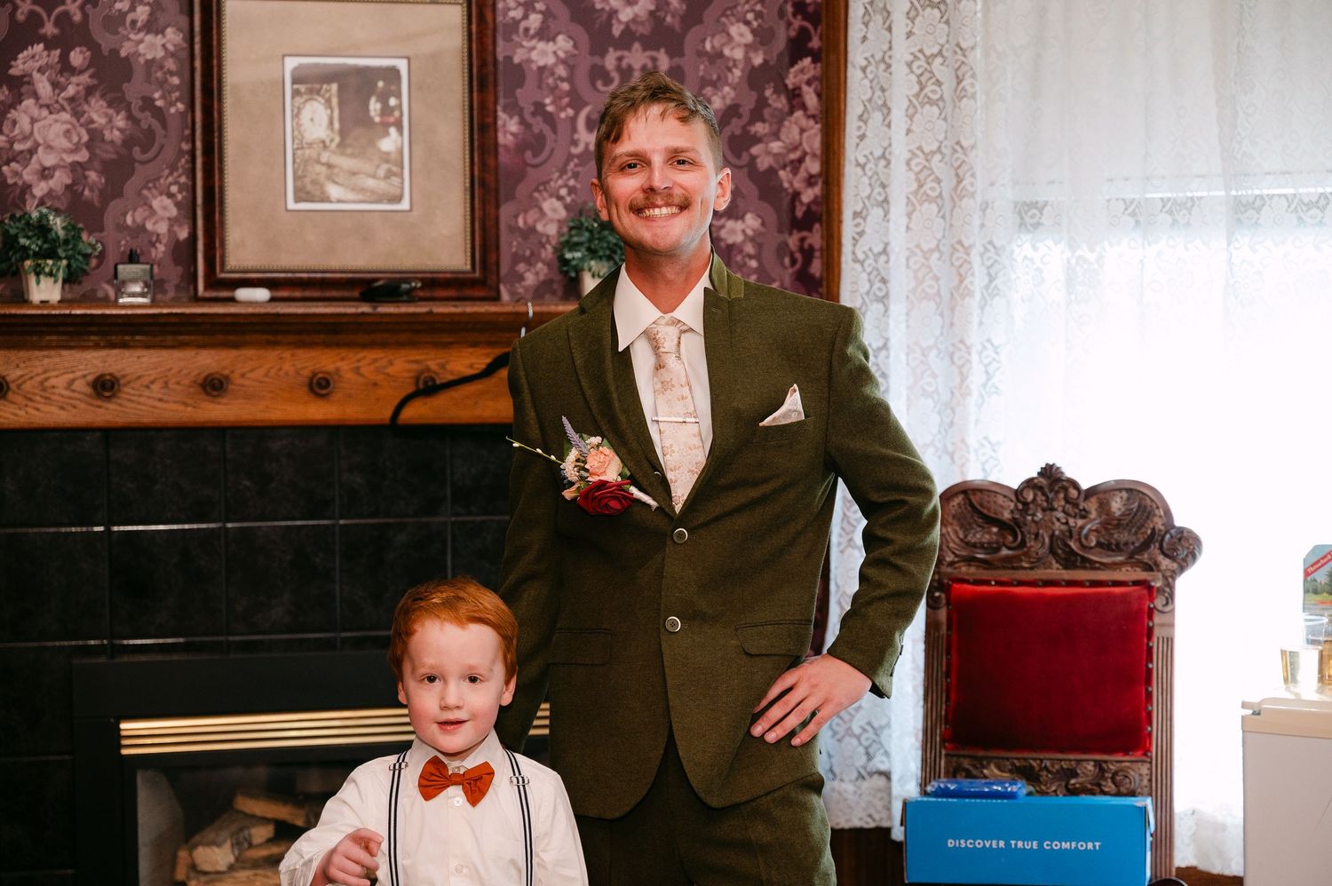 Central MN Wedding Photos | Man in green suit with boutonniere poses with young red-haired boy in bow tie near fireplace at wedding.