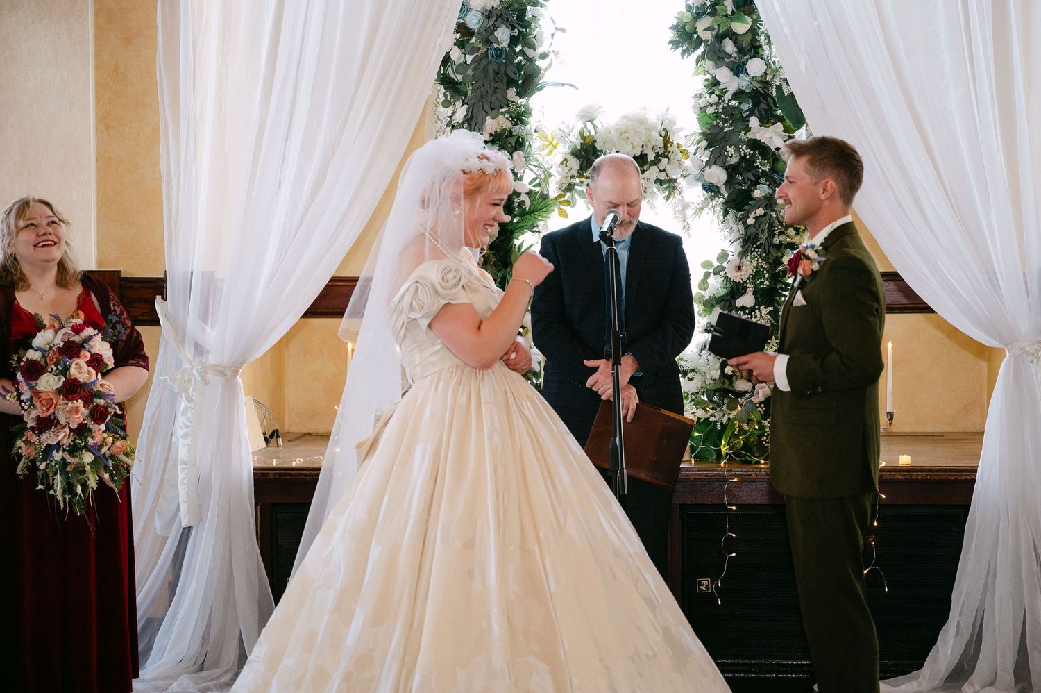 Central MN Wedding Photos | Bride and groom exchange vows at altar adorned with white drapes and Christmas trees in ornate venue.
