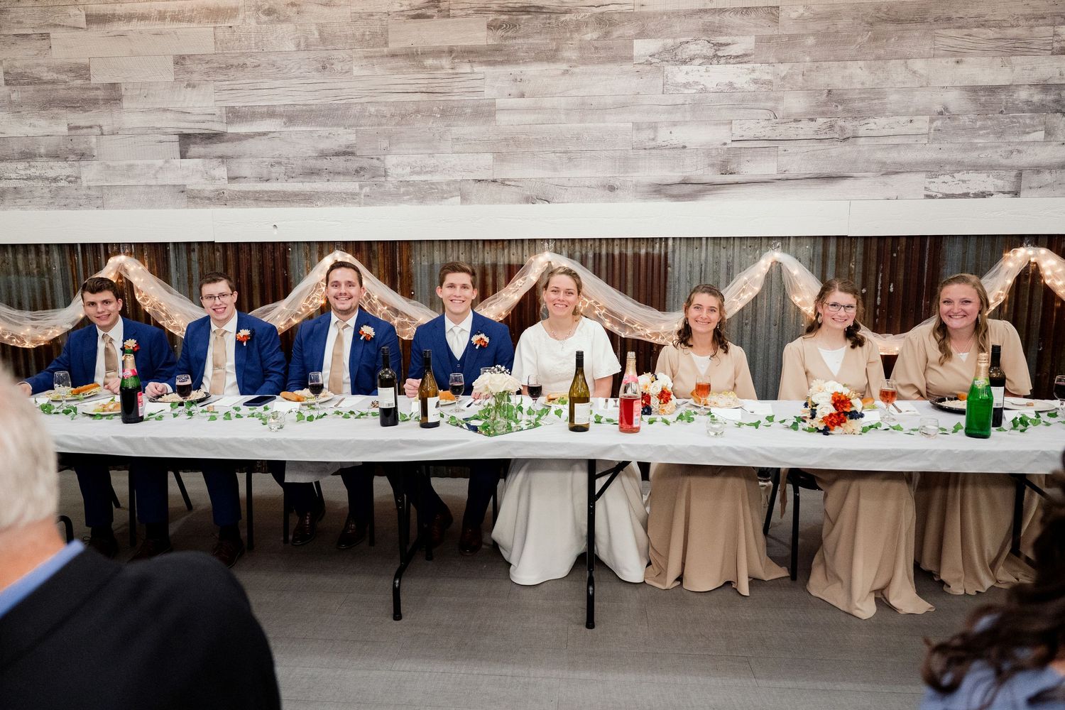 Wedding party seated at long reception table showing joyful expressions with arms raised in celebration.
