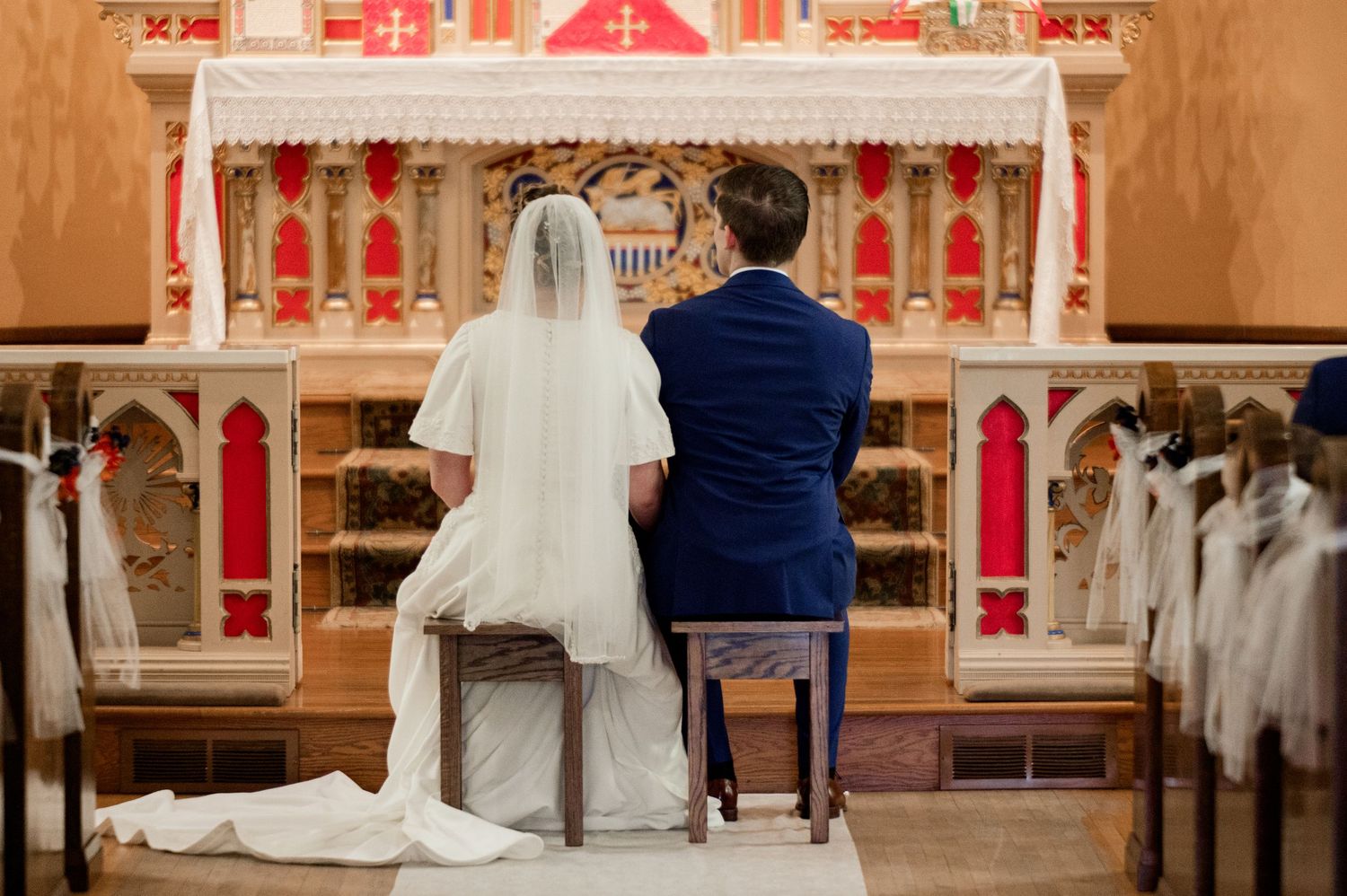 View from behind of couple seated at ornate church altar with red and gold decor during wedding ceremony.