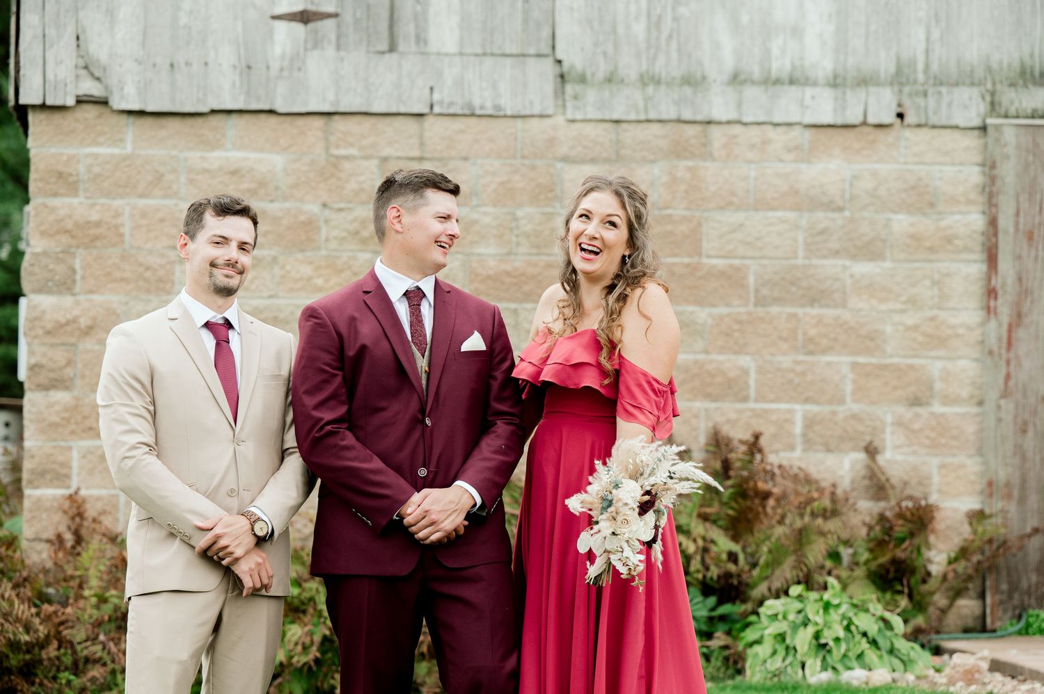 Wedding party members in burgundy and tan attire pose against stone wall at rustic venue.