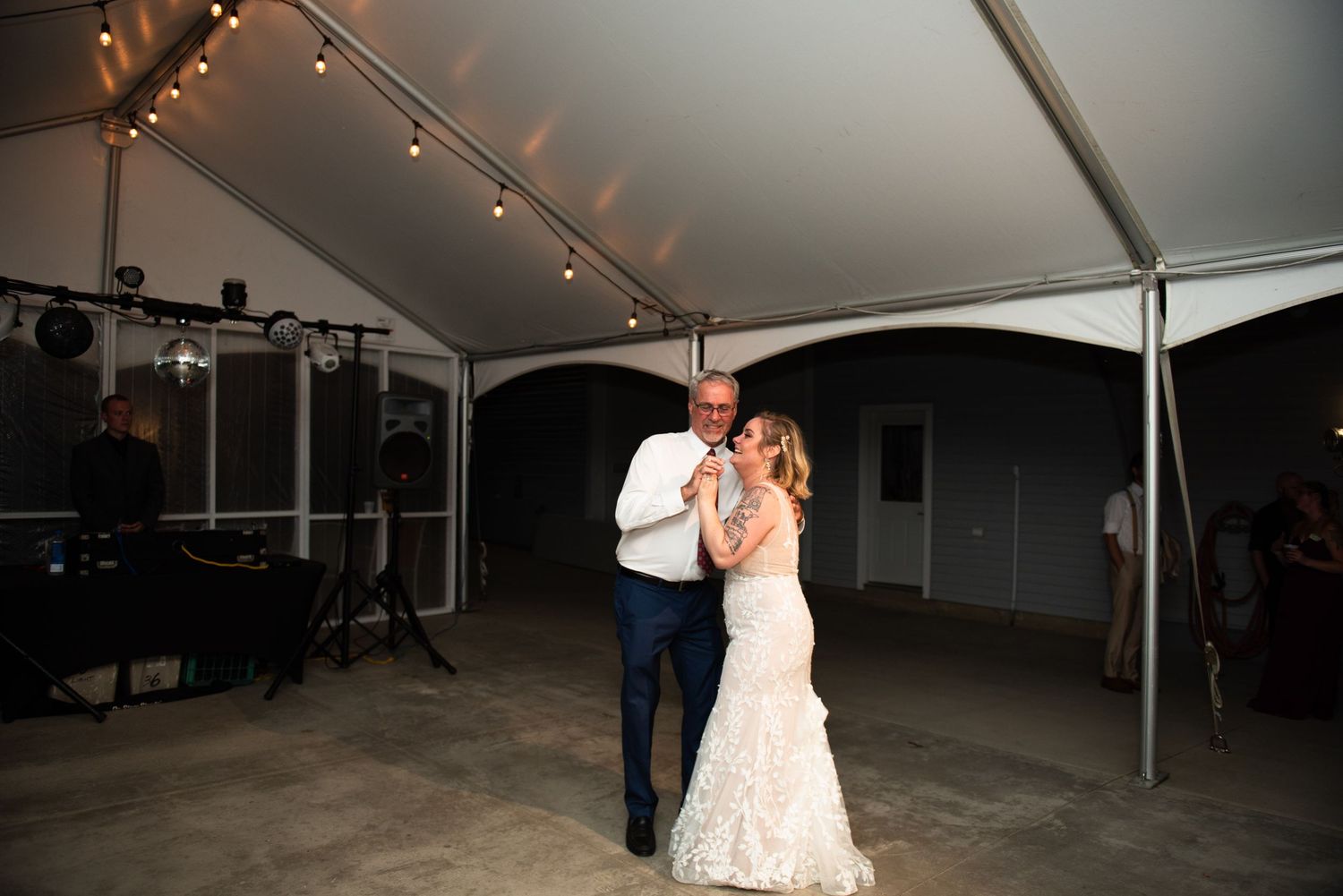 A romantic slow dance under a white tent with string lights at a wedding reception.