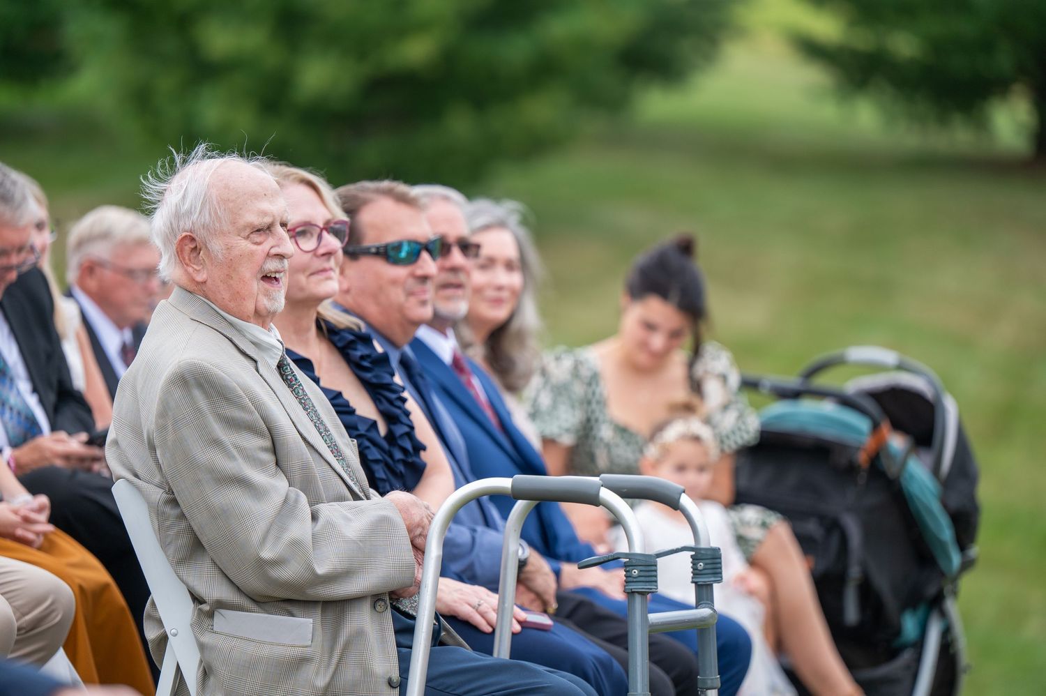 Wedding guests seated outdoors watching ceremony on sunny day with natural greenery in background.