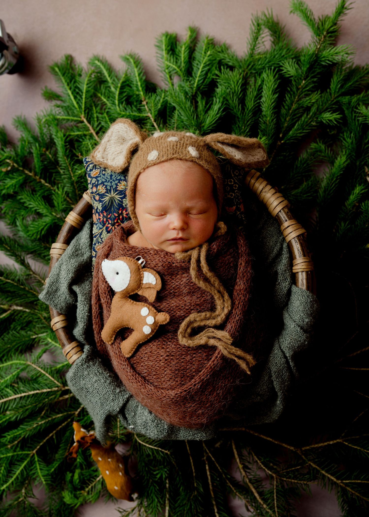 Baby sleeping peacefully on evergreen branches wearing a knit cap with woodland animal accents.