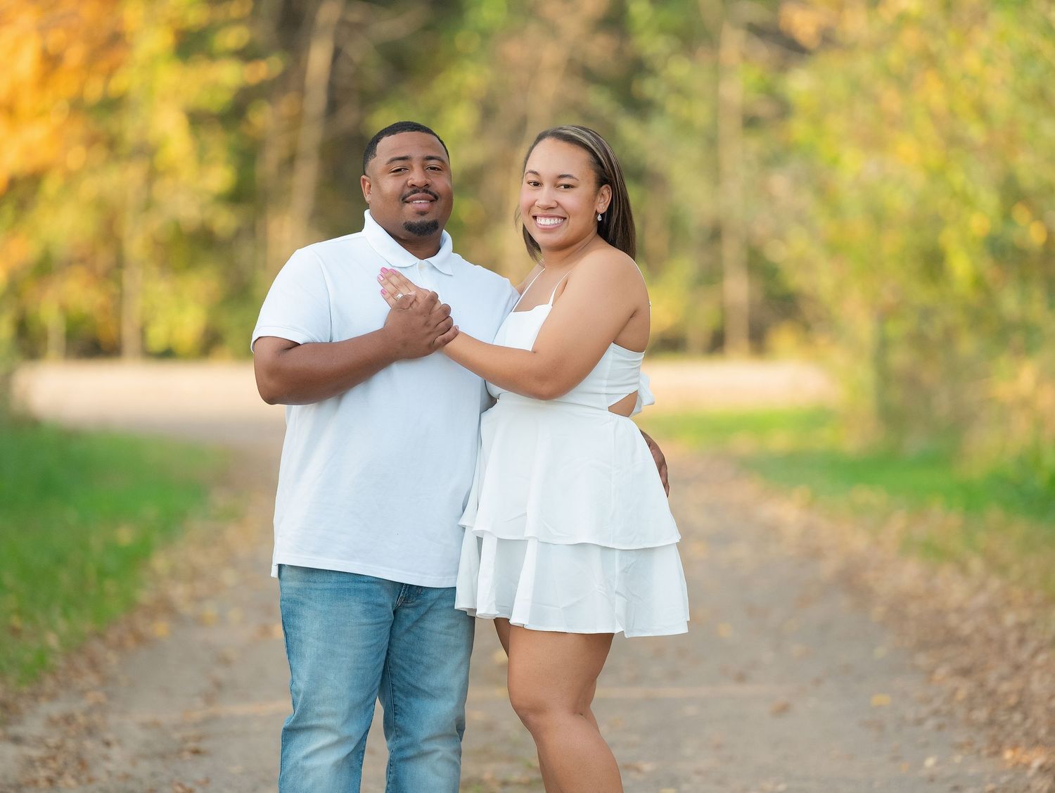 A couple in coordinated white and denim outfits embrace on a scenic autumn path surrounded by golden foliage.